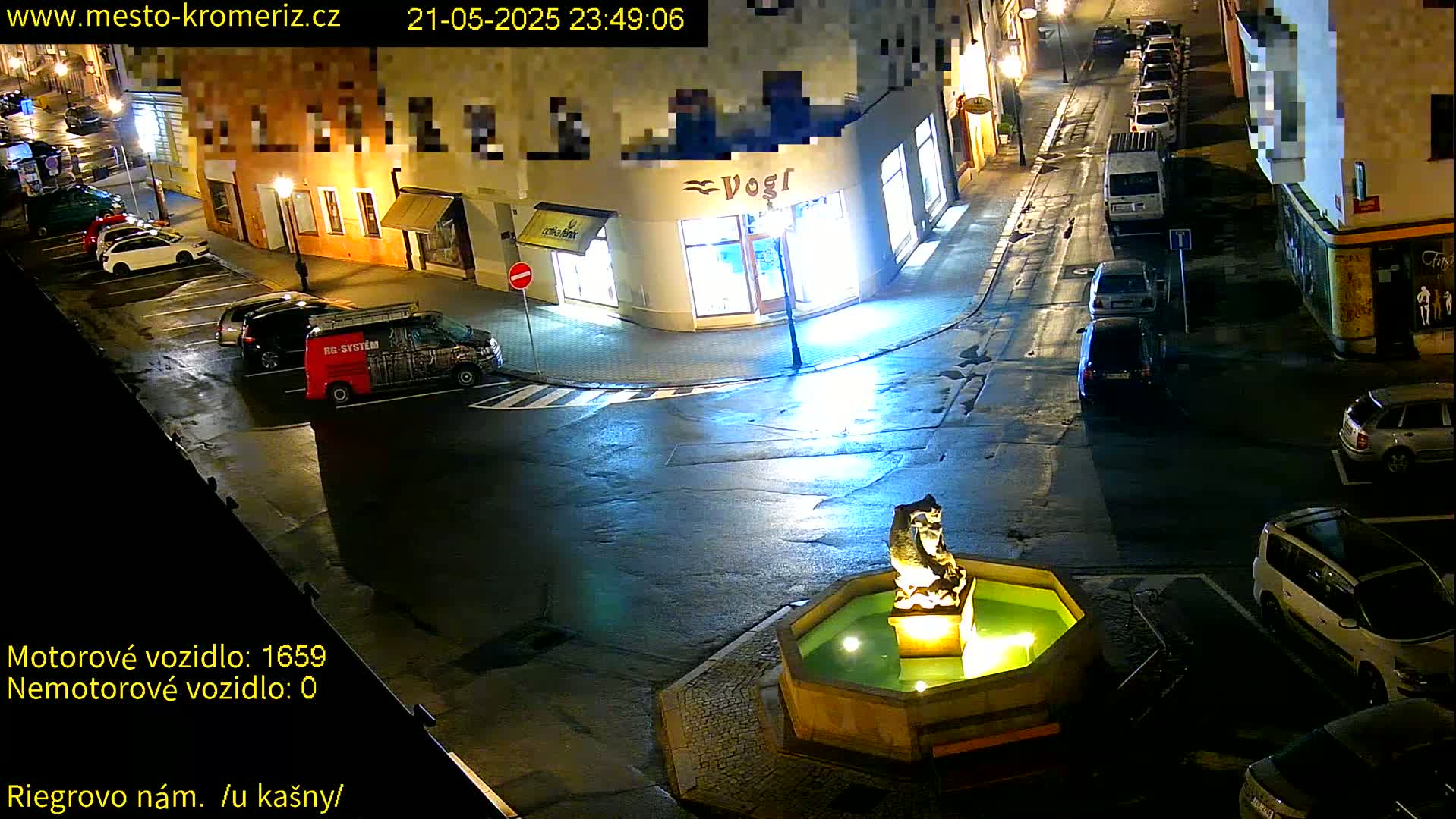 A nighttime street scene shows several parked cars surrounding a lit fountain with a stone sculpture.