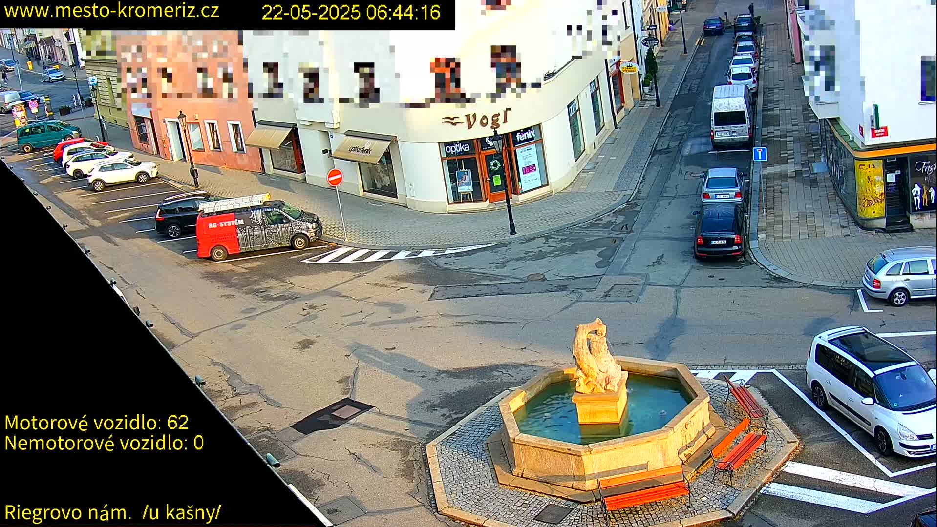 A town square with a fountain and several parked cars, under sunny conditions.