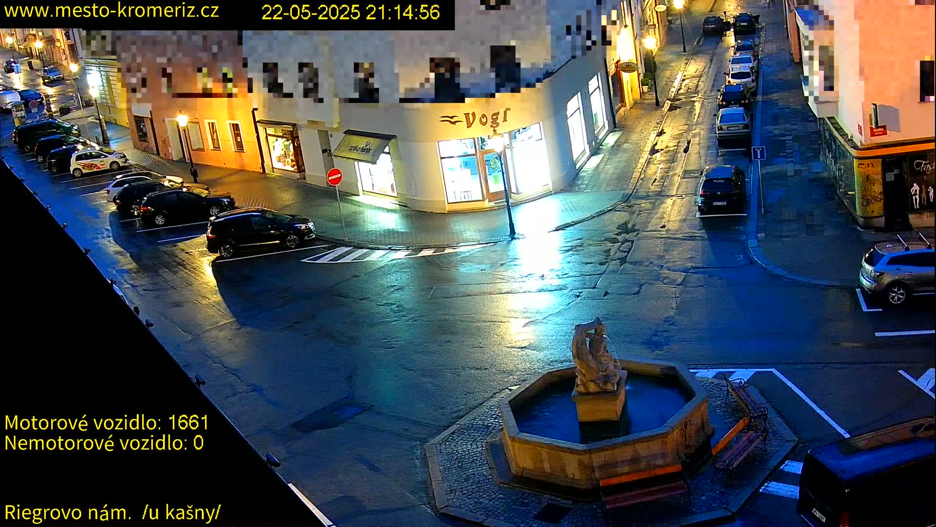A wet, nighttime street scene shows a fountain in a town square with parked cars along the sides of the buildings.