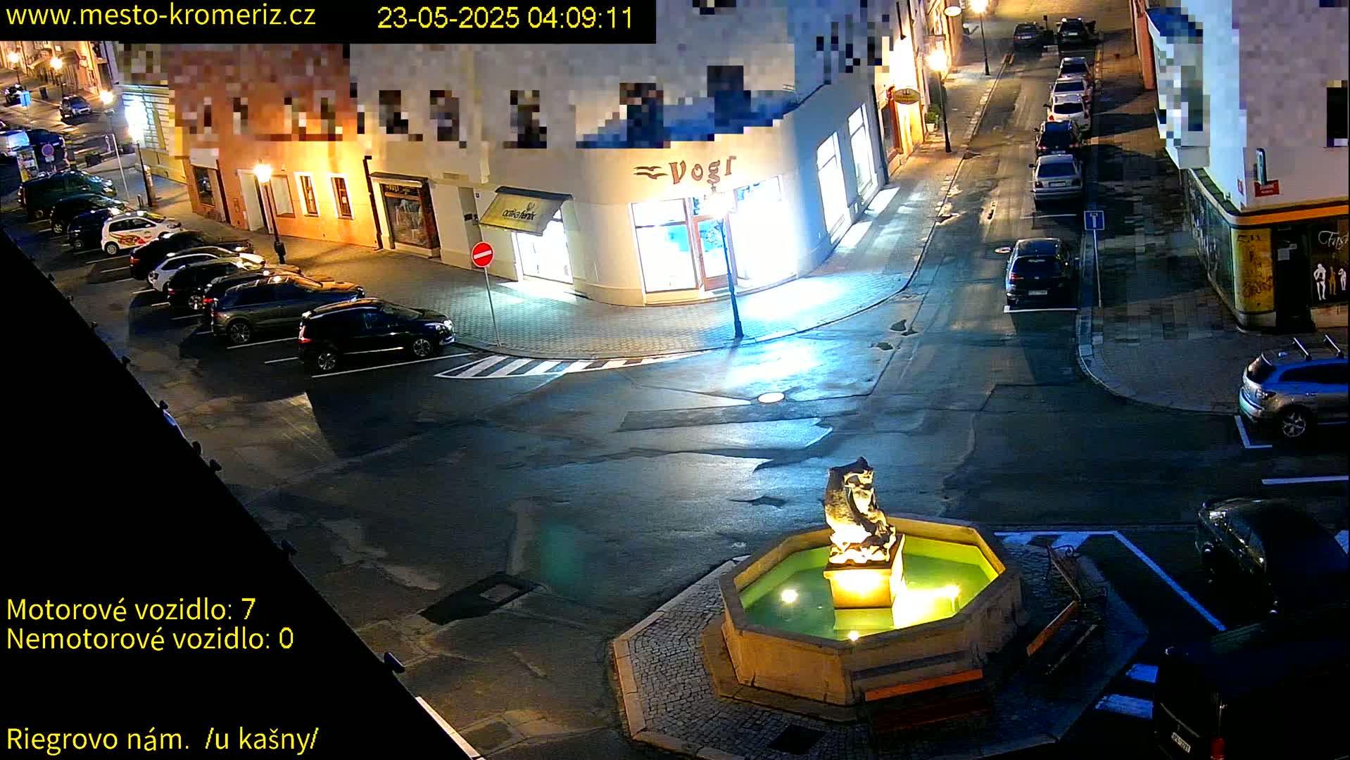 At night, a town square with parked cars surrounds a fountain featuring a statue, illuminated under dim streetlights.