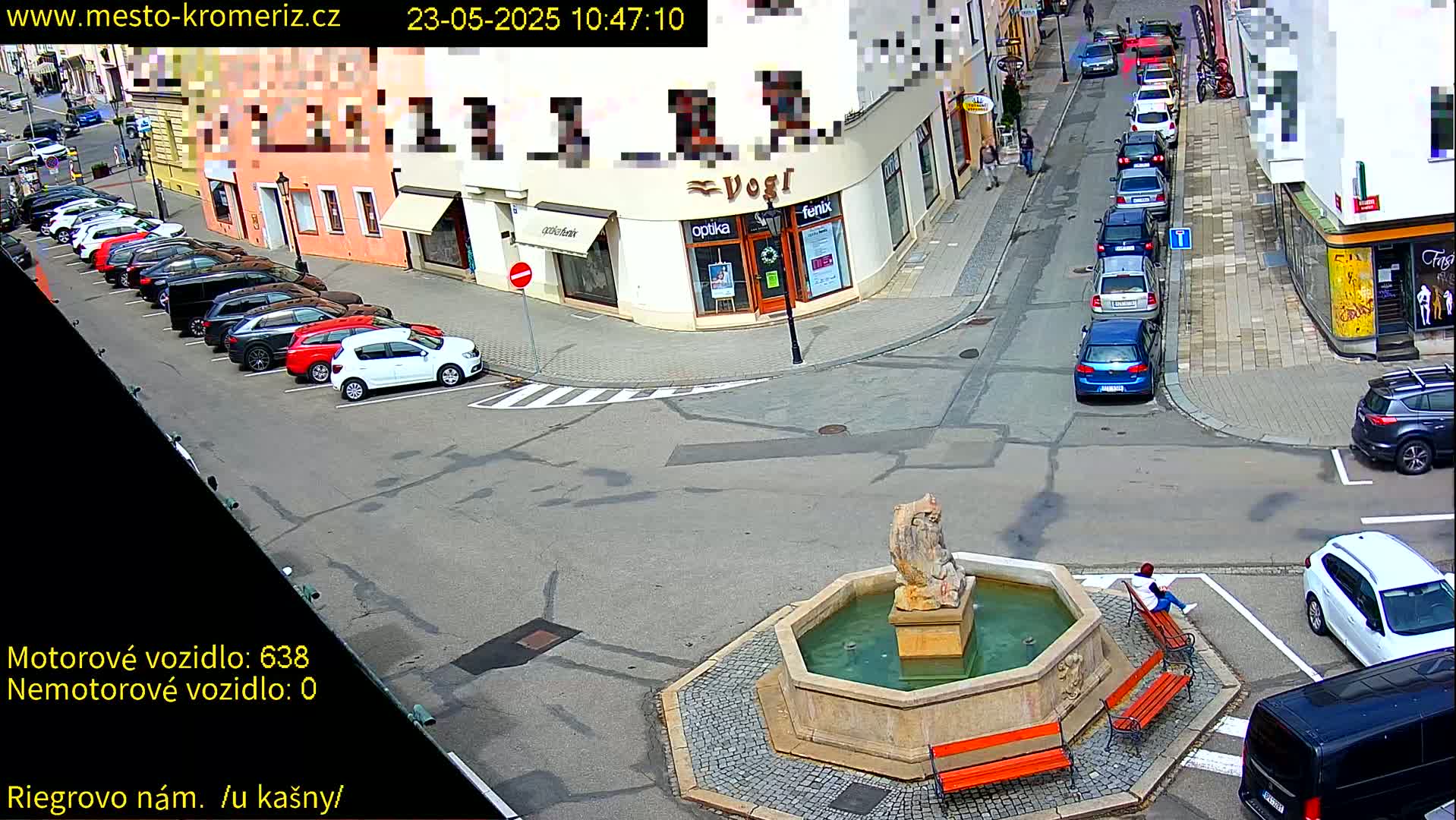 A sunny town square features a stone fountain with a statue, several parked cars lining the street, and a person sitting on a bench.