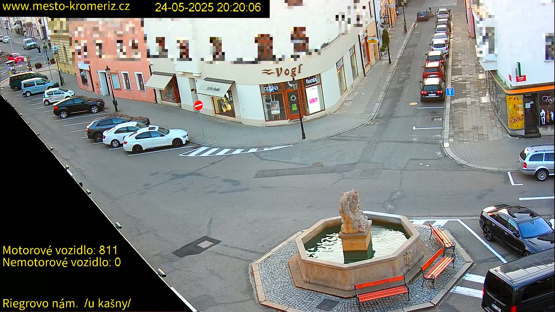 Several cars are parked along the street and around a stone fountain in a town square under clear evening conditions.