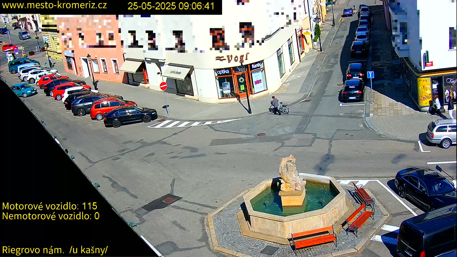 A sunny day shows a town square with a fountain in the center, surrounded by parked cars and buildings, and a person riding a bicycle.