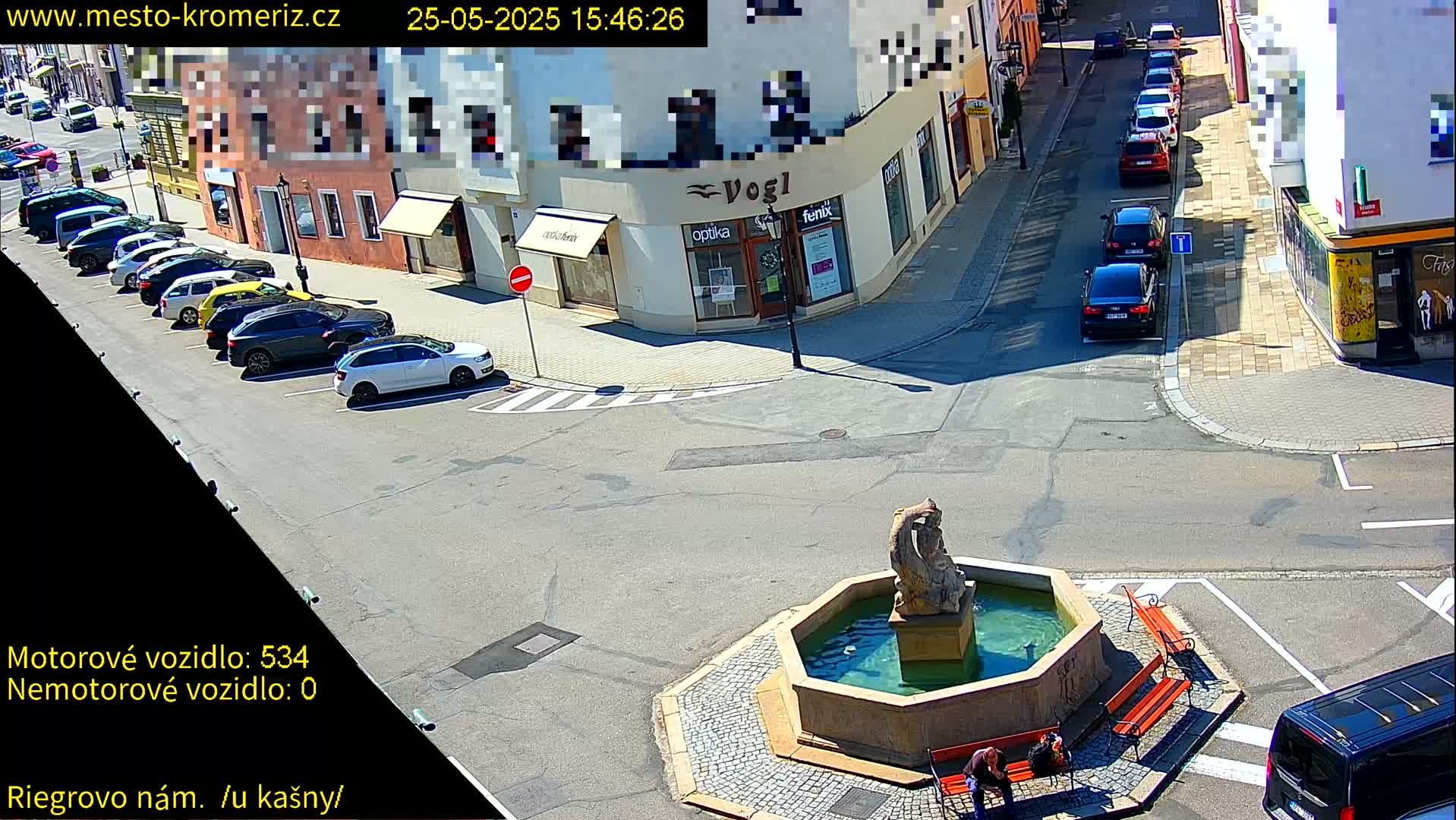 A sunny town square with a fountain in the center, surrounded by parked cars and buildings, with a few pedestrians near the fountain.