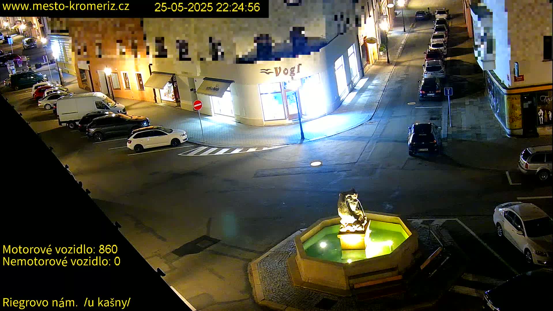 A nighttime street scene shows a town square with parked cars, a fountain with a statue, and buildings along the streets.