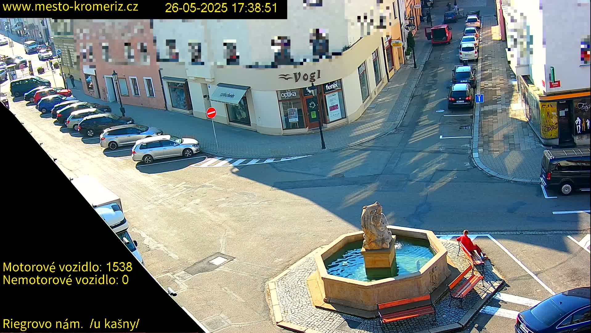 A sunny street scene shows a town square with a fountain, several parked cars, and a person sitting on a bench.