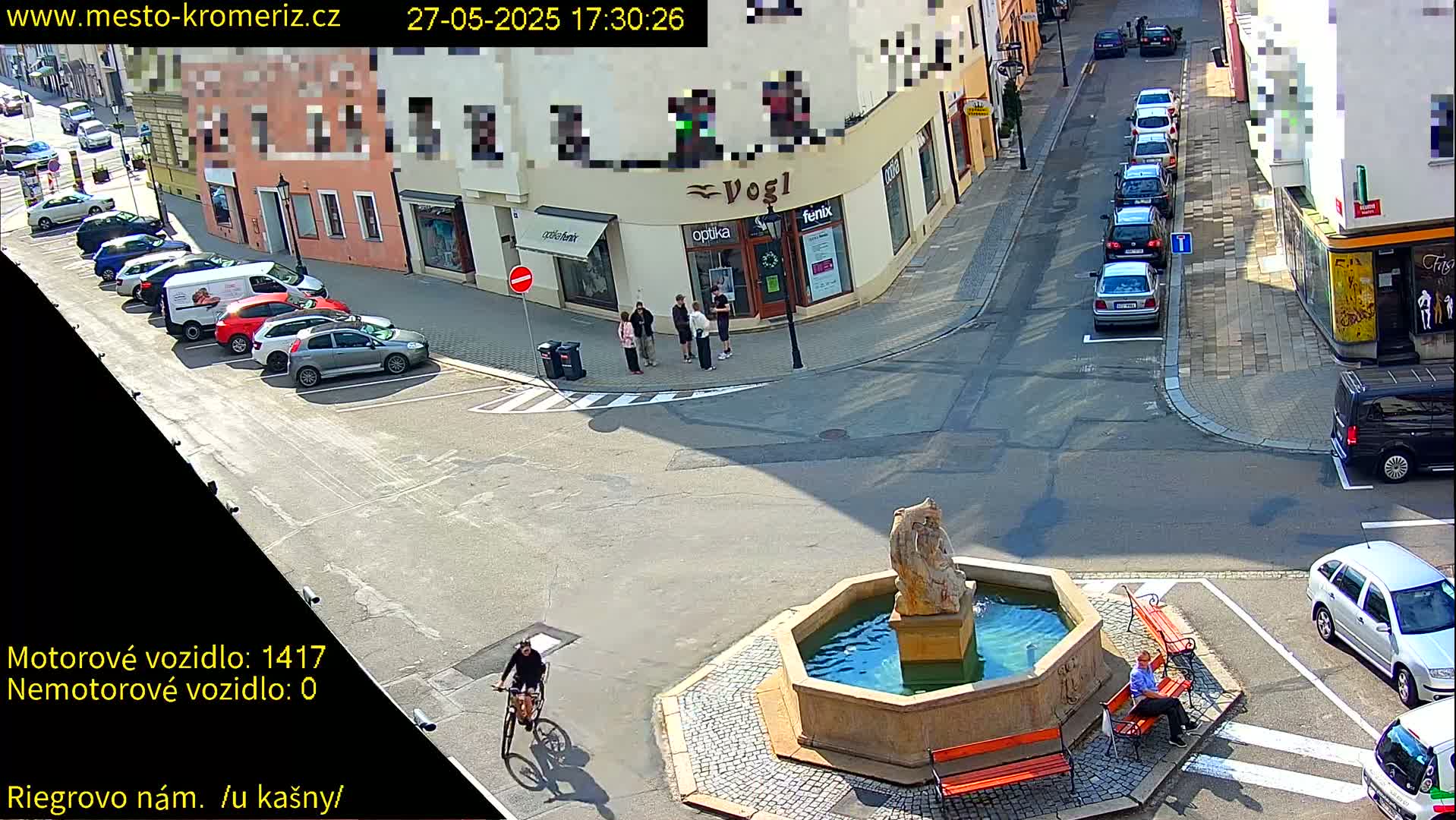 A sunny day shows a town square with a fountain, several parked cars, a person on a bicycle, and a person sitting on a bench.