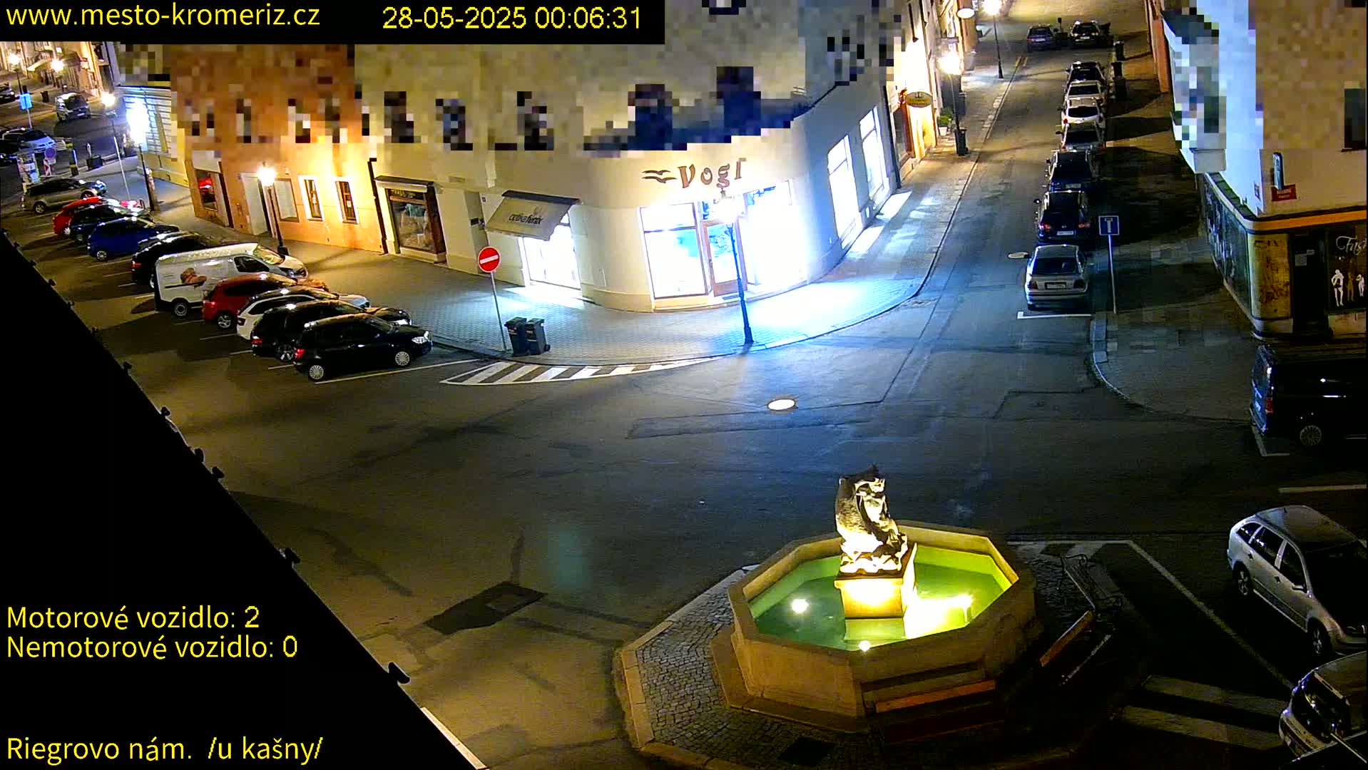 A nighttime street scene shows several parked cars lining the sides of a street that converges at a fountain with a statue in the center.