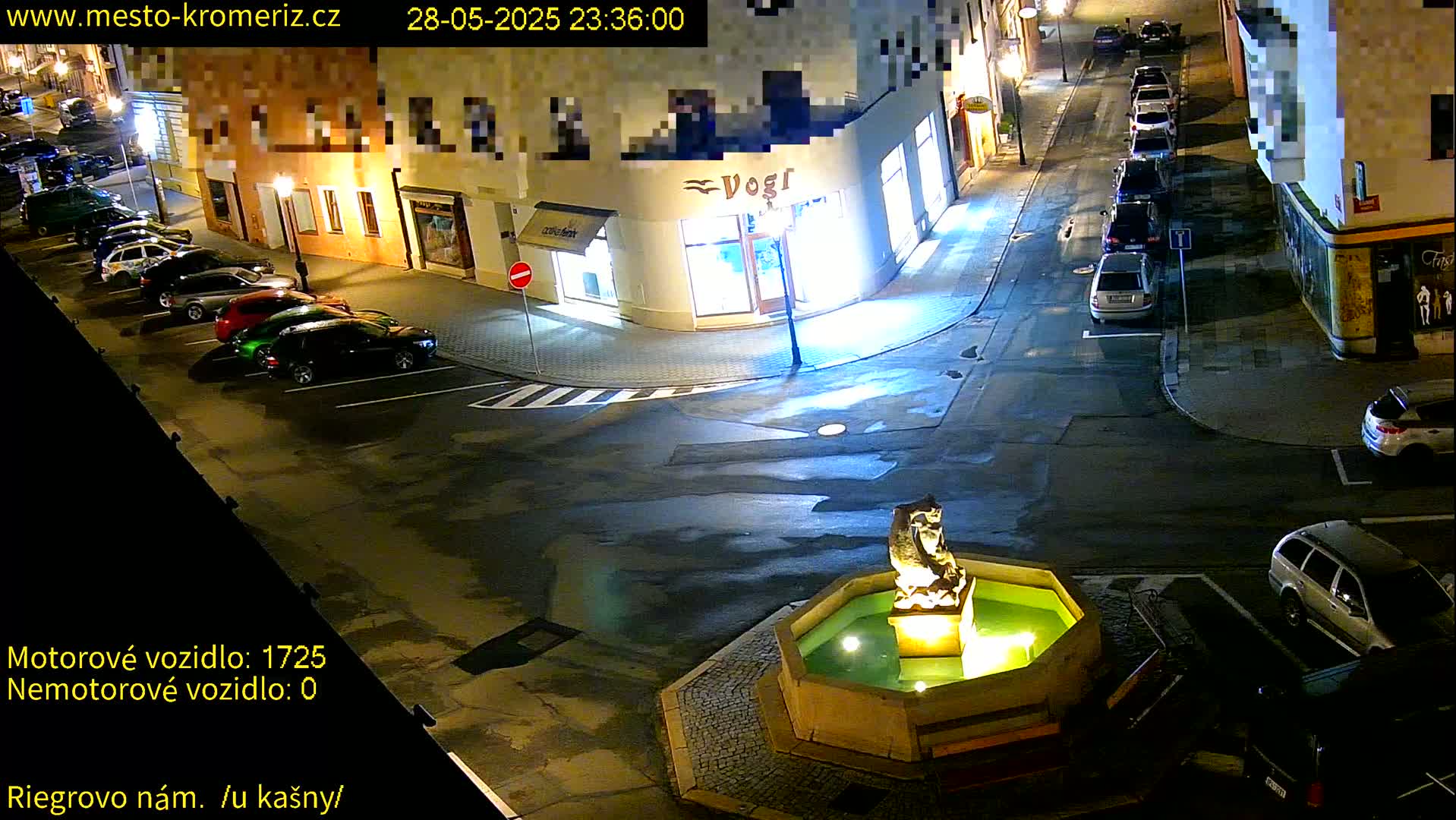 A nighttime street scene shows several cars parked along the curb near a fountain with a statue, illuminated by streetlights.