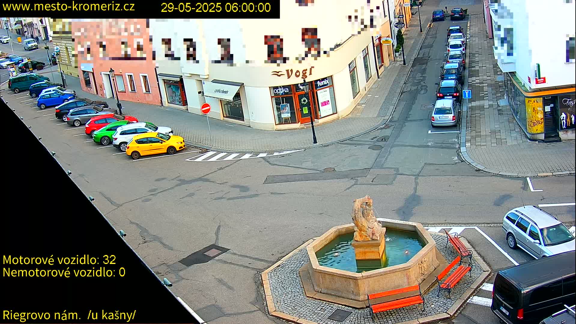 A town square with a stone fountain, several parked cars, and buildings lining the streets on a sunny day.