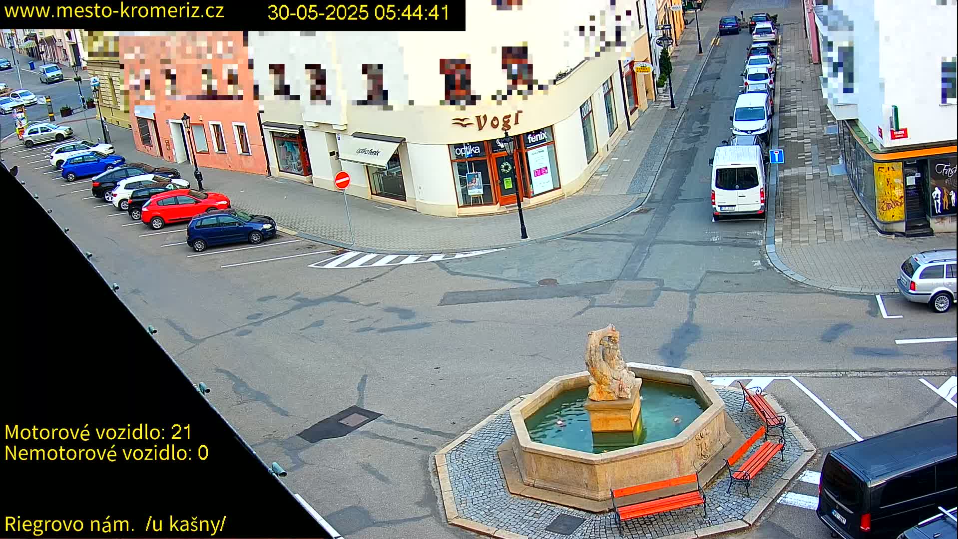 A town square with a fountain at its center, several parked cars lining the streets, and a few cars driving by under a clear sky.
