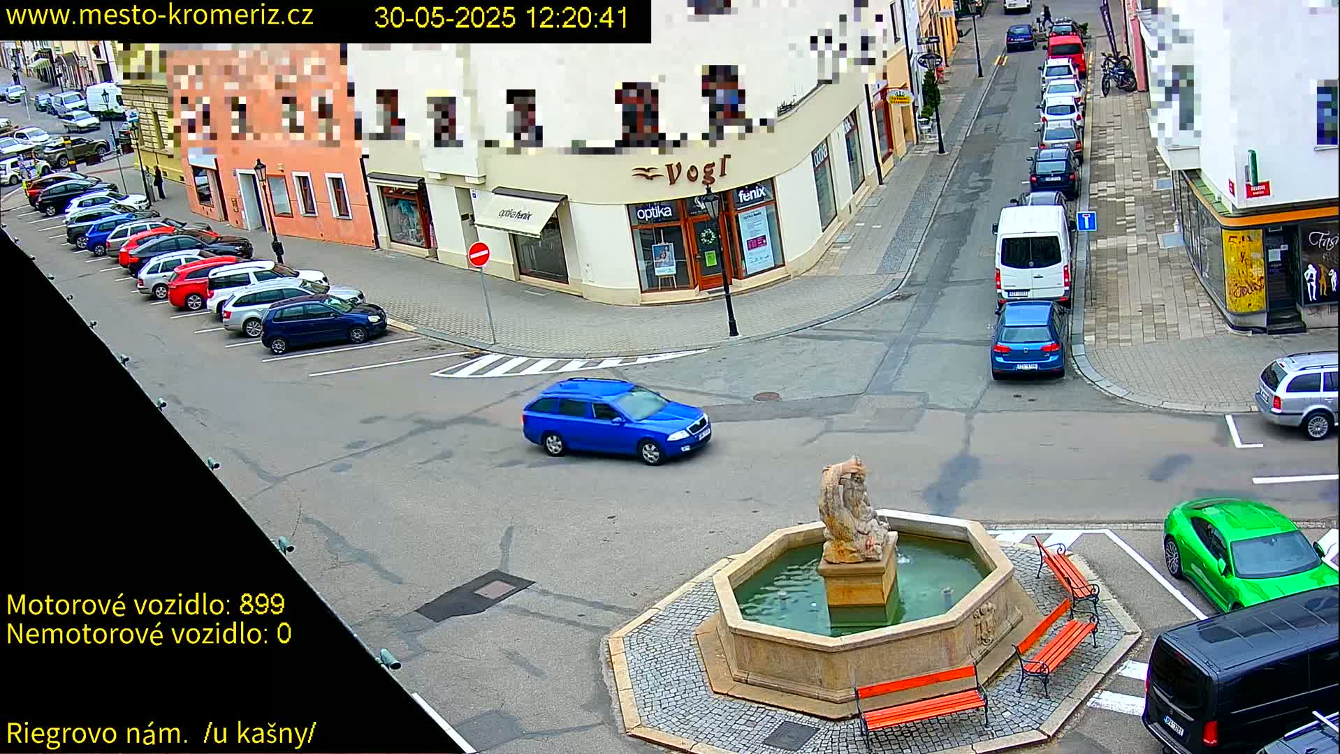 A blue car drives past a stone fountain in a town square with parked cars and buildings lining the streets on a sunny day.