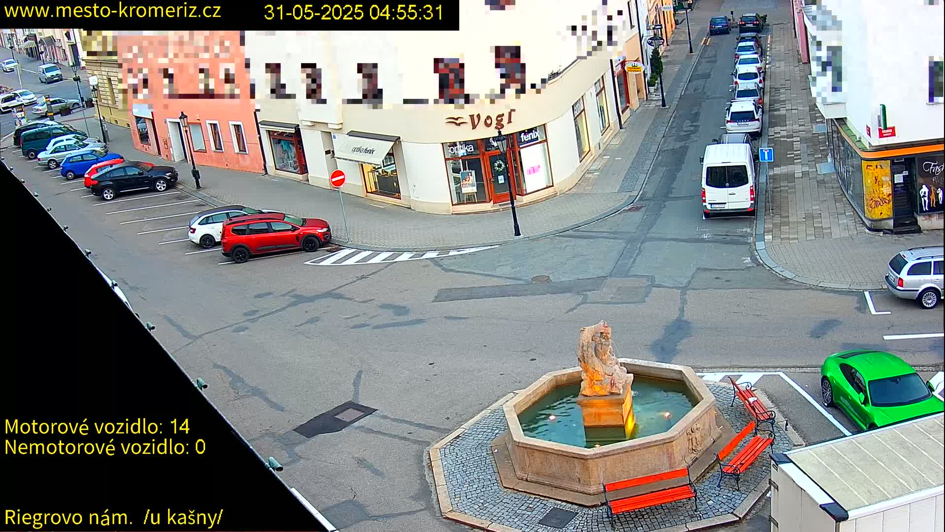 A street scene shows several parked cars surrounding a stone fountain with a statue, under a clear sky.