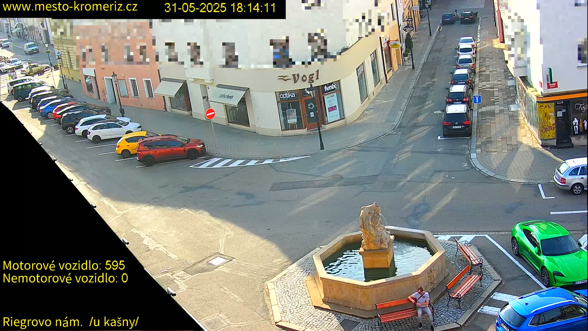 A sunny town square with a stone fountain, several parked cars, and a person sitting on a bench.