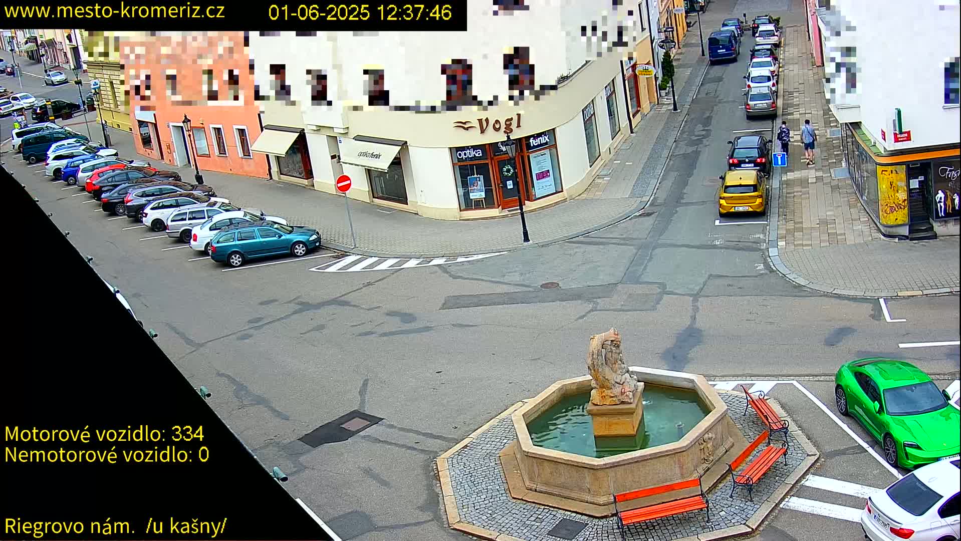 A sunny day shows a town square with a stone fountain, several parked cars, and a few pedestrians.