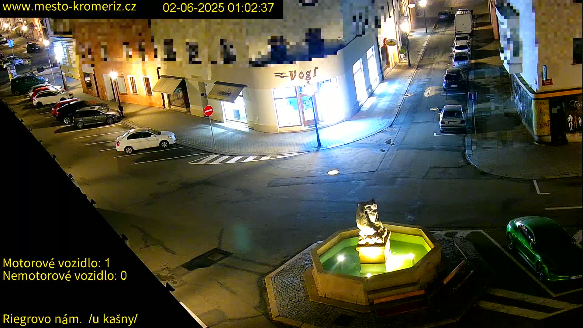 At night, a town square with several parked cars surrounds a fountain featuring a stone statue.