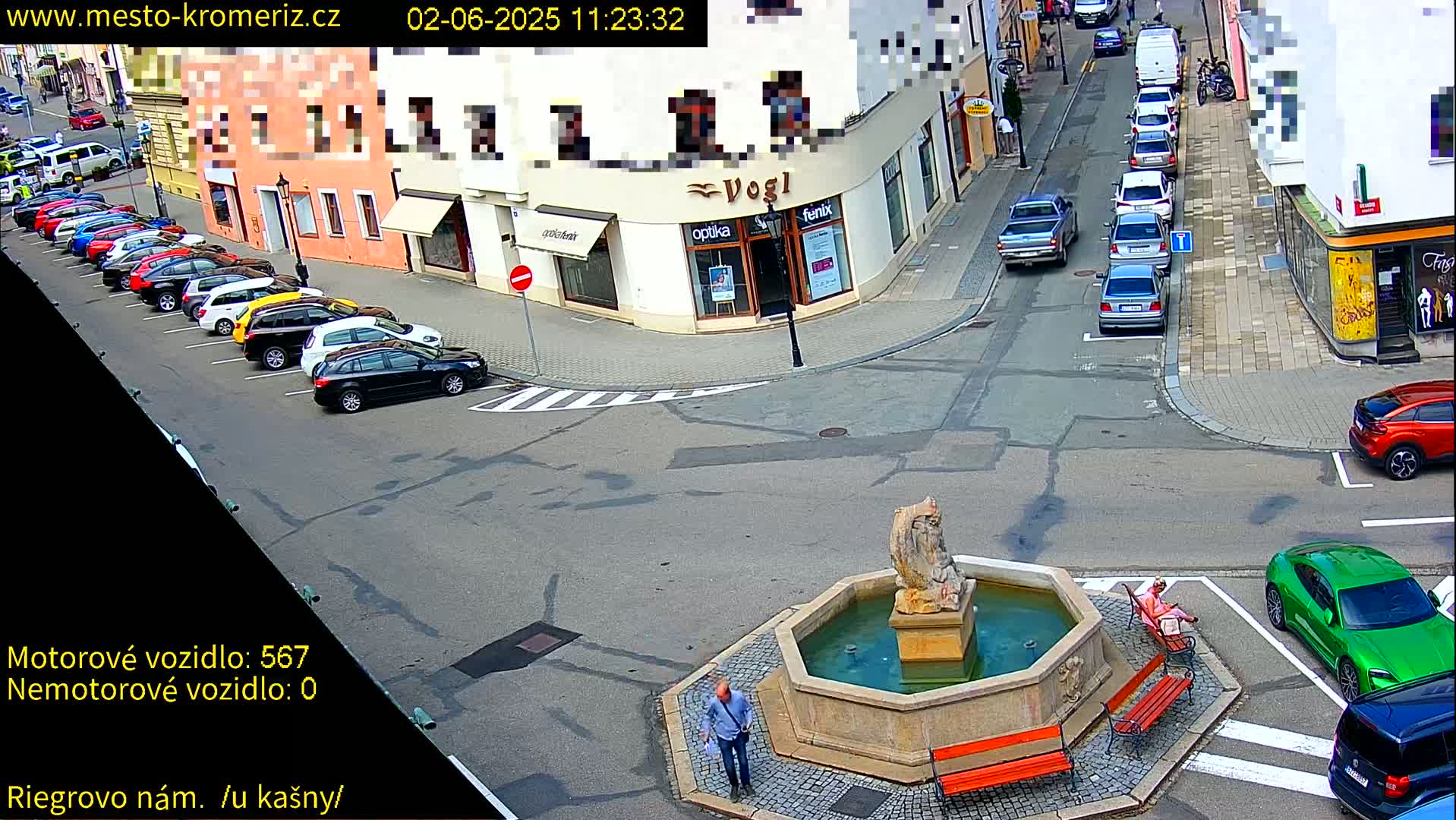 A sunny day shows a town square with a fountain, several parked cars, and a person standing near the fountain.