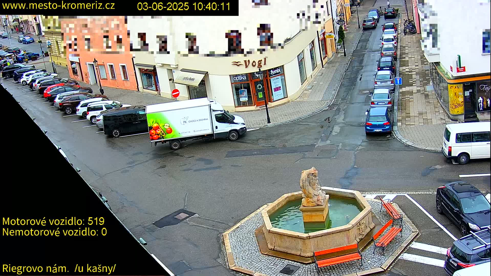 A town square with a stone fountain, several parked cars, and a delivery truck, viewed on a slightly overcast day.