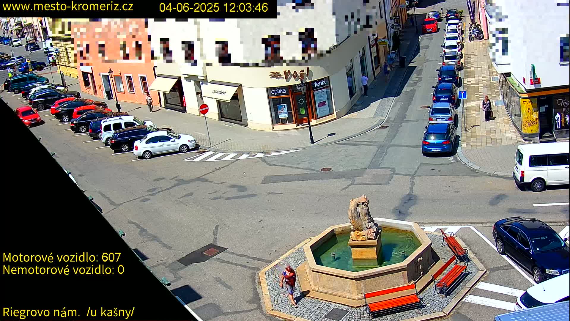 A sunny street scene shows a town square with a stone fountain, several parked cars, and a few pedestrians; some cars are parked along a street.