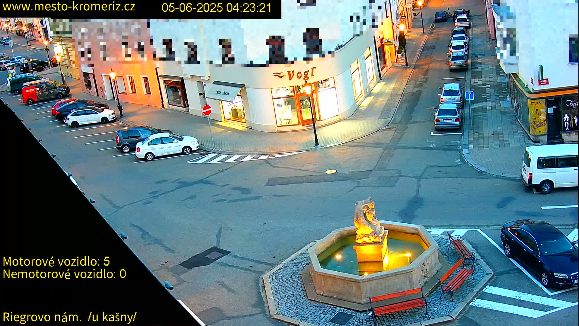 A town square at dusk, featuring a stone fountain, several parked cars, and some buildings with lit windows.