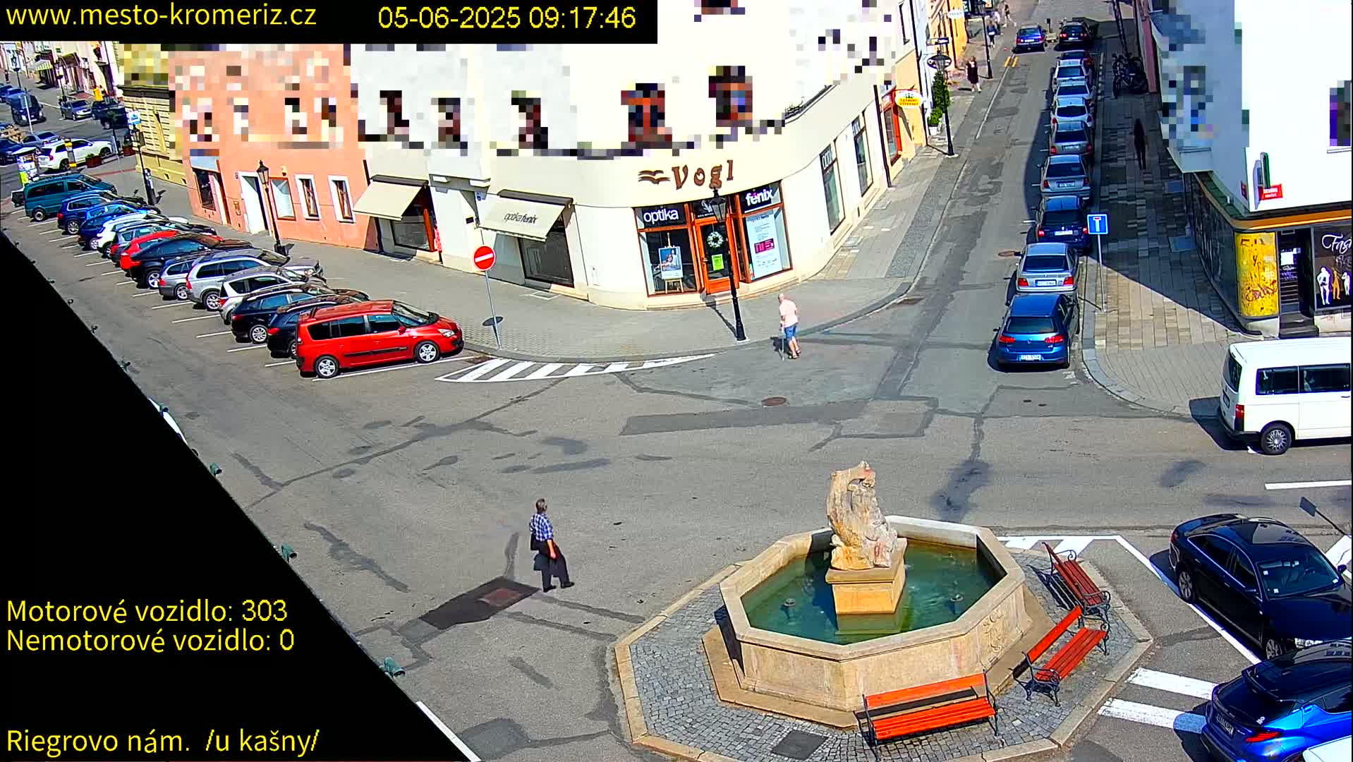 A sunny town square with a fountain at its center, surrounded by parked cars and pedestrians.