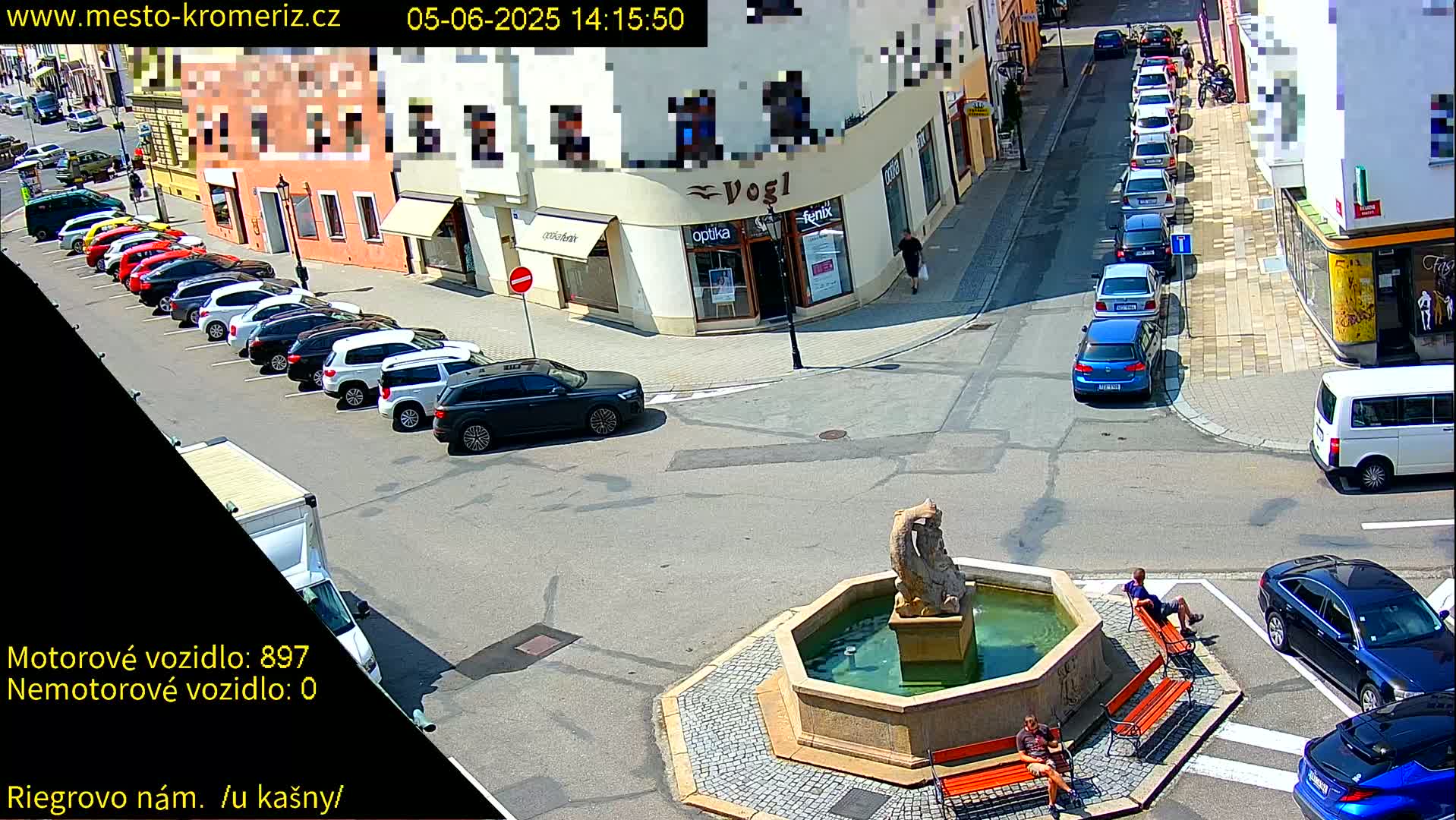 A town square with a fountain, several parked cars, and a few people sitting on benches on a sunny day.