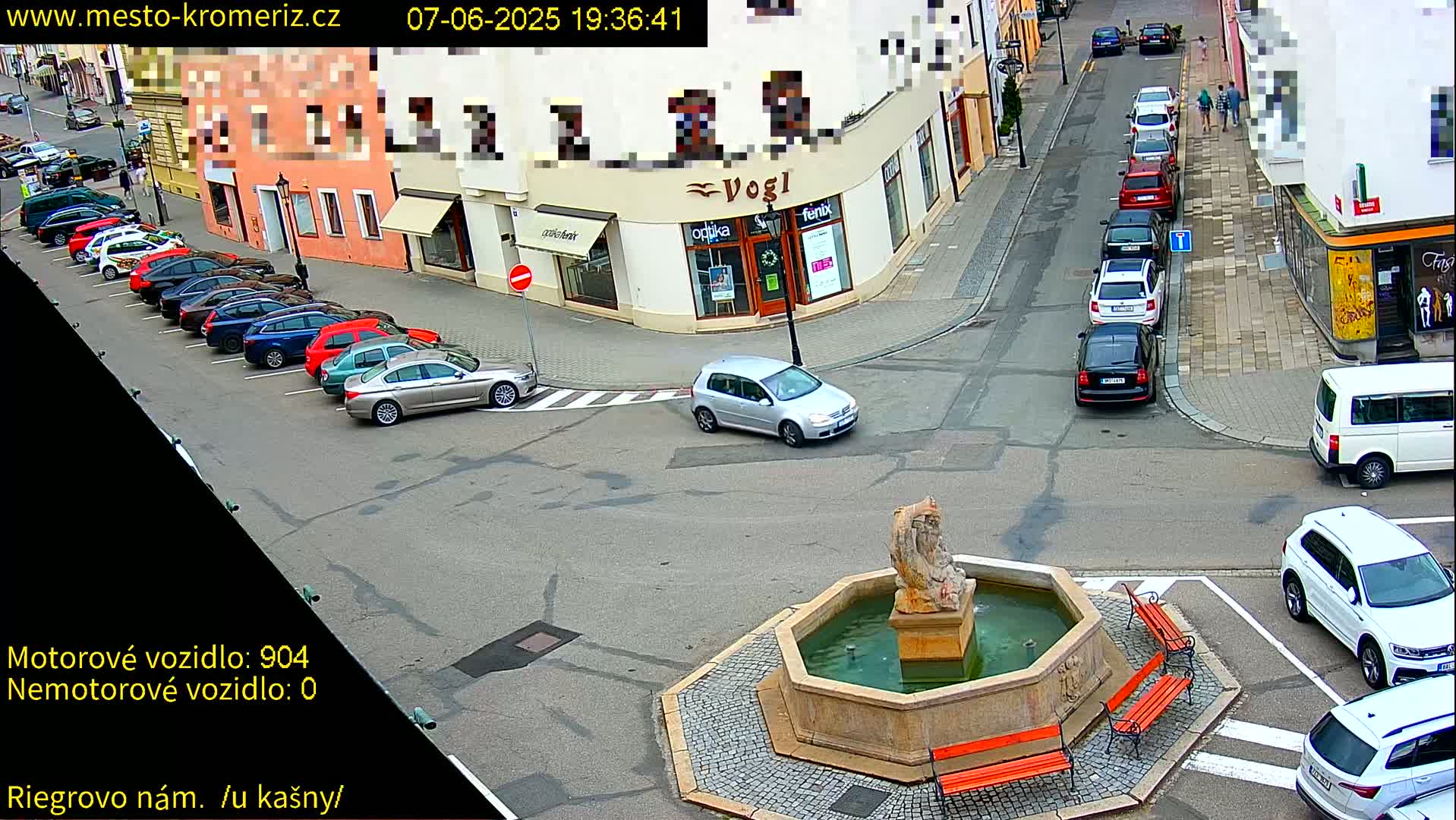 A town square with a fountain at its center, surrounded by parked cars and buildings, under daylight conditions.