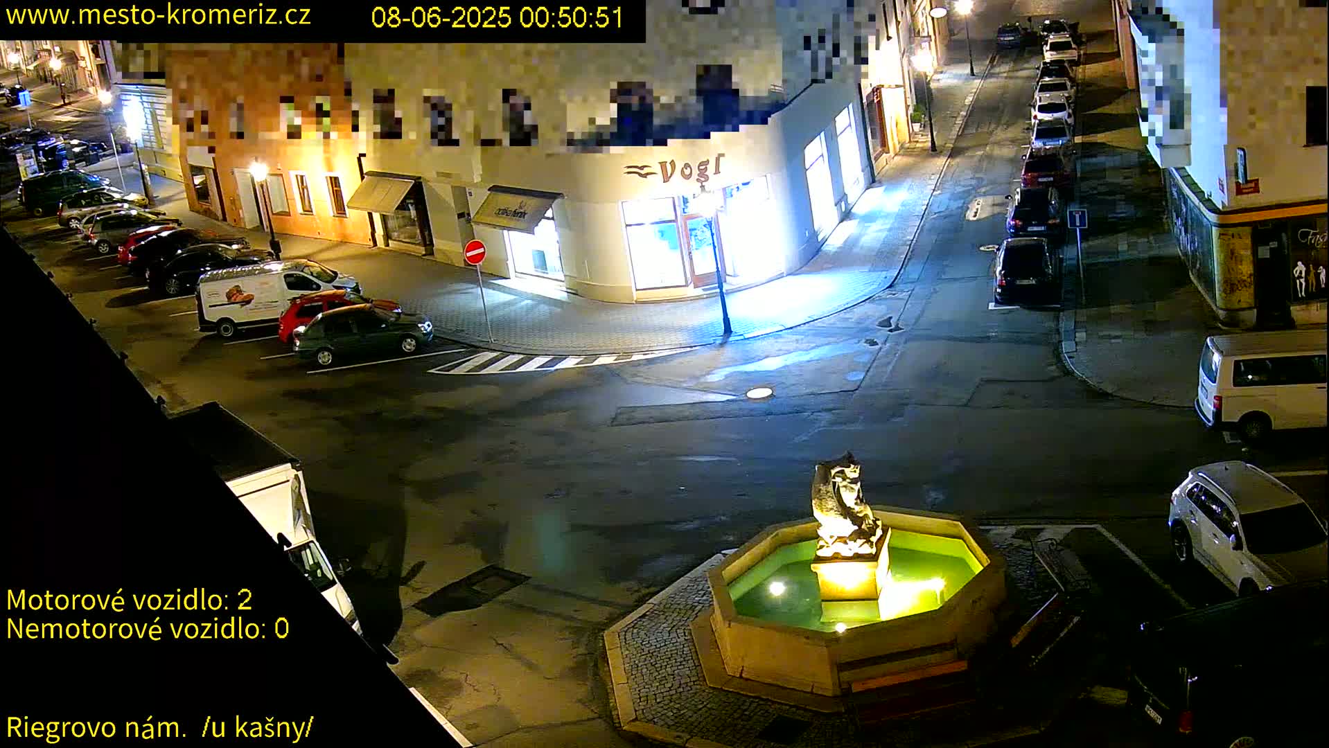 A nighttime street scene shows a town square with a fountain, several parked cars, and some buildings.