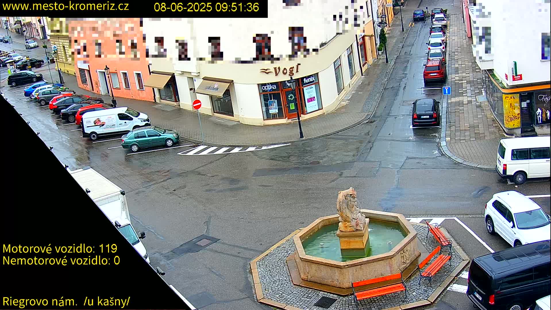 A wet town square with a stone fountain, surrounded by parked cars and buildings.