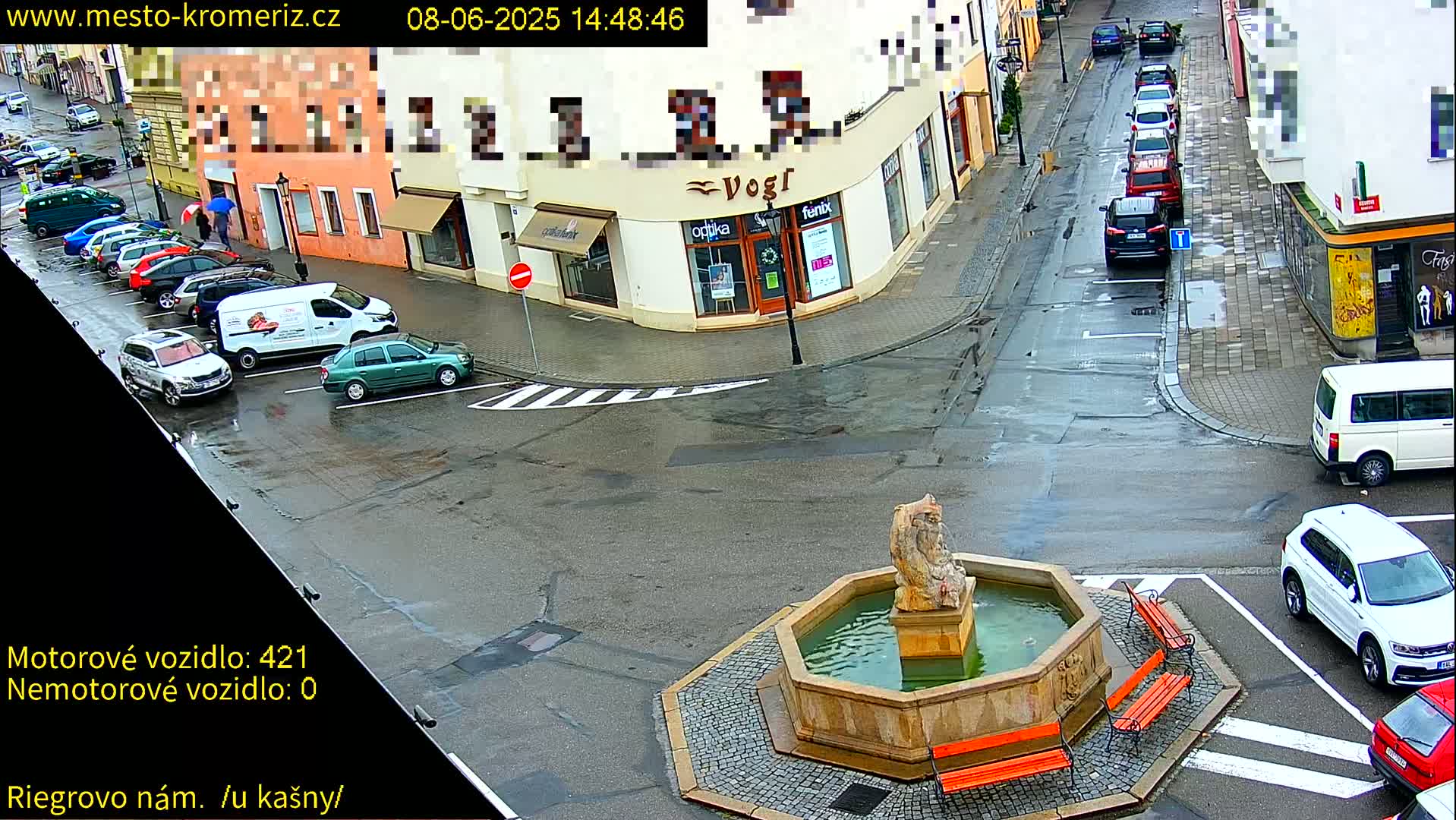 A wet town square with parked cars and a stone fountain is viewed from above.