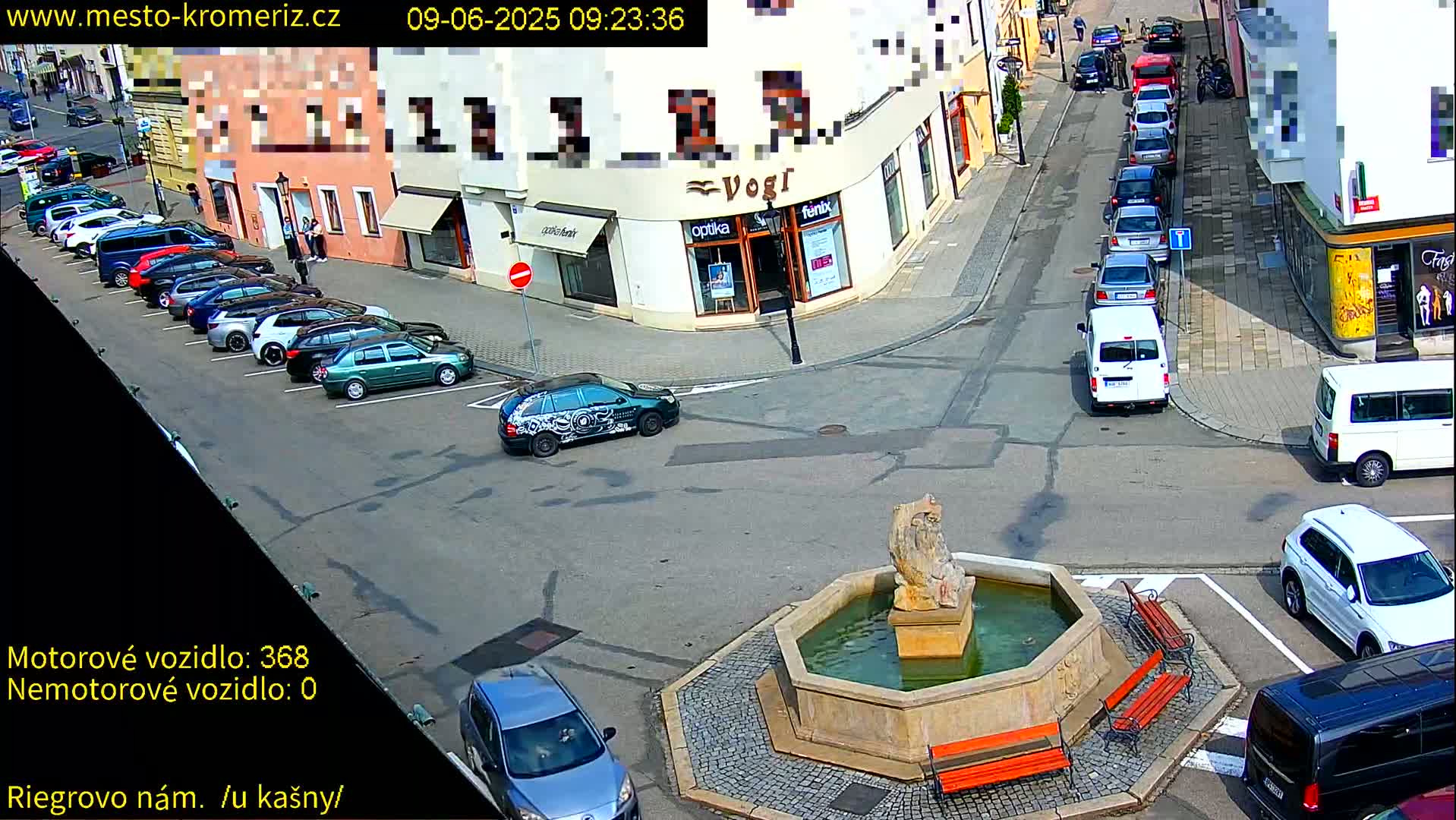 A sunny town square shows numerous parked cars around a stone fountain with benches, and several cars are driving on the surrounding streets.