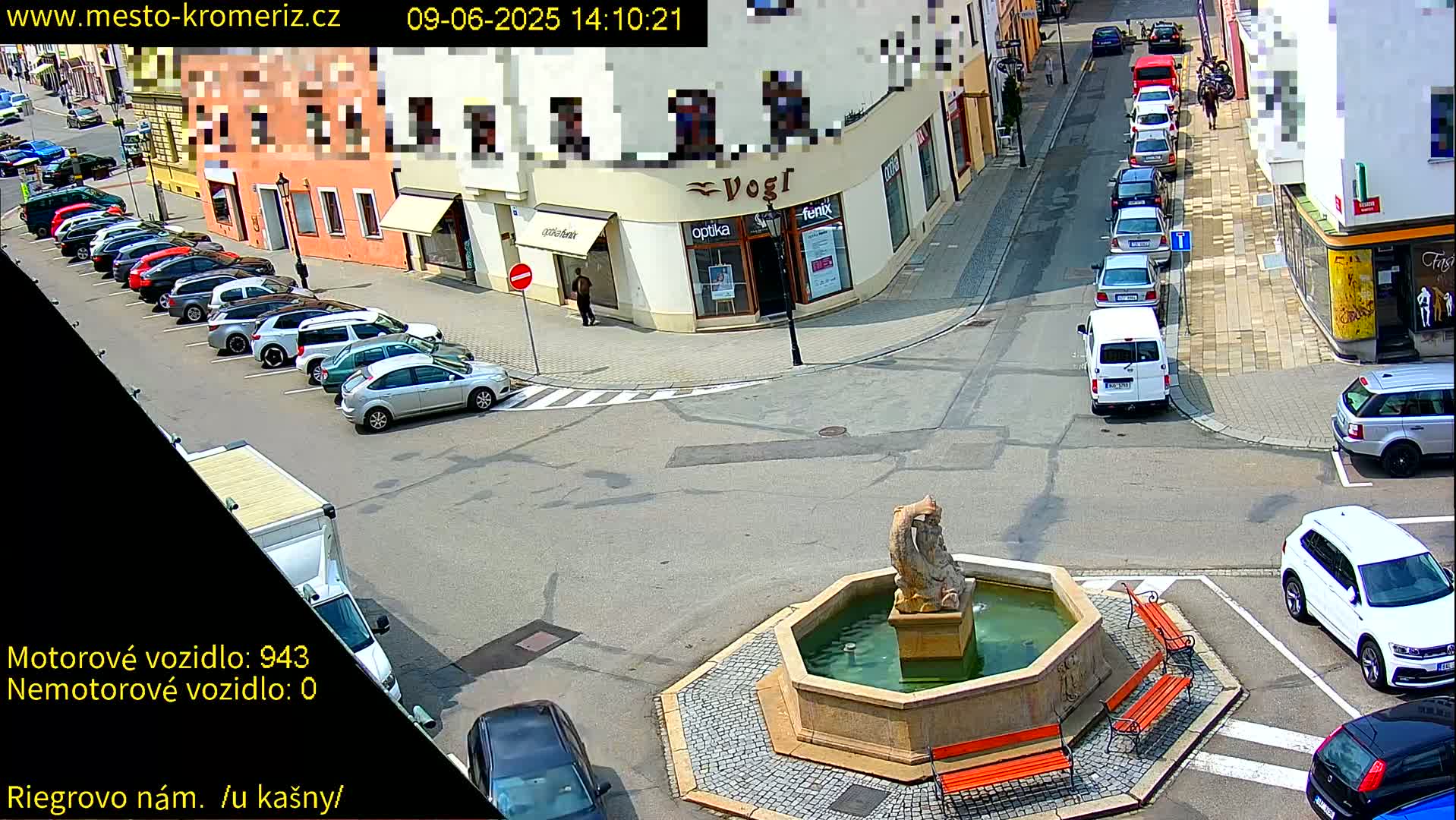 A sunny town square with a fountain and several parked cars, some cars are driving on the streets surrounding the square.
