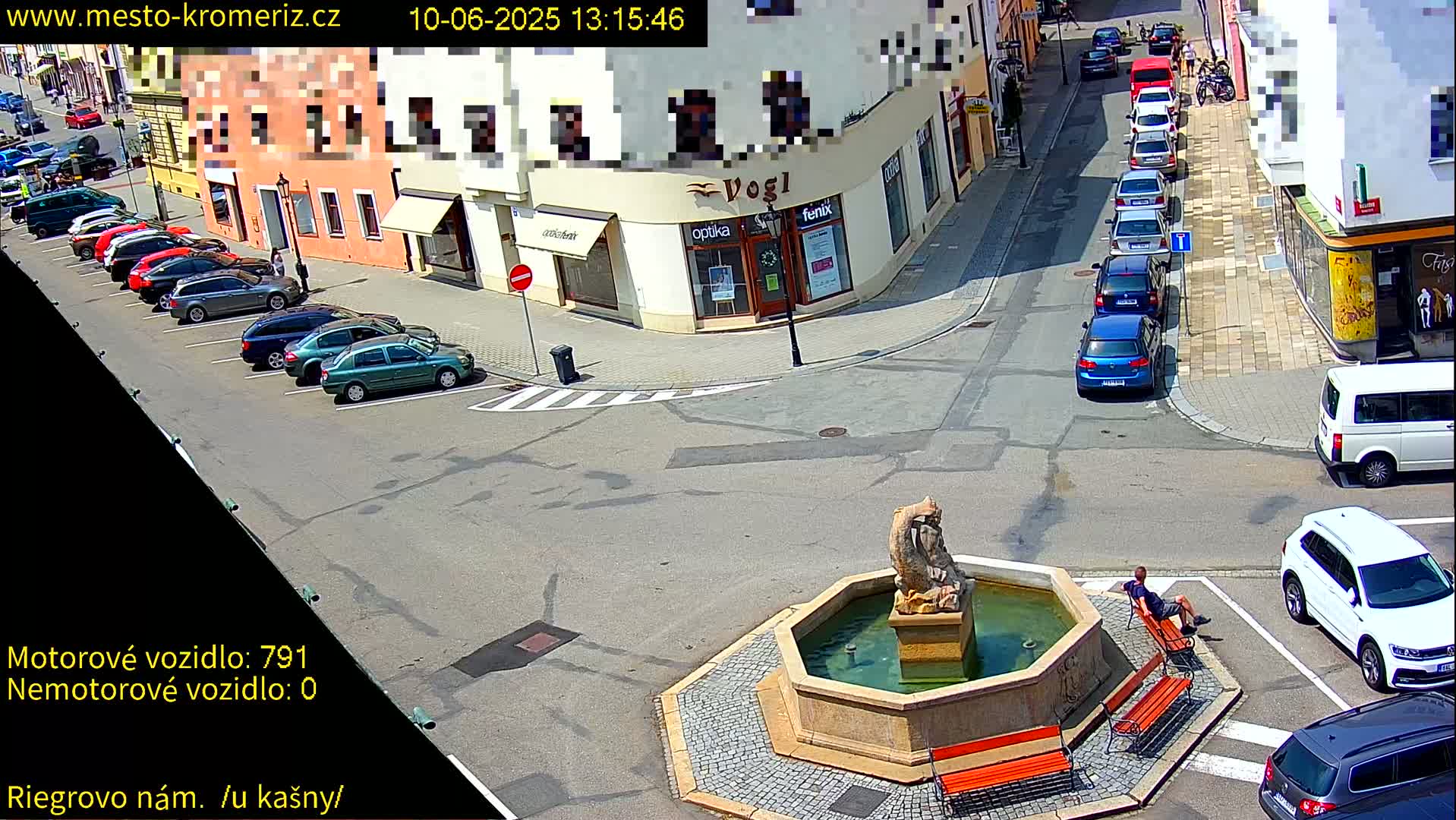 A sunny day scene shows a town square with a fountain, several parked cars, and a person sitting on a bench.