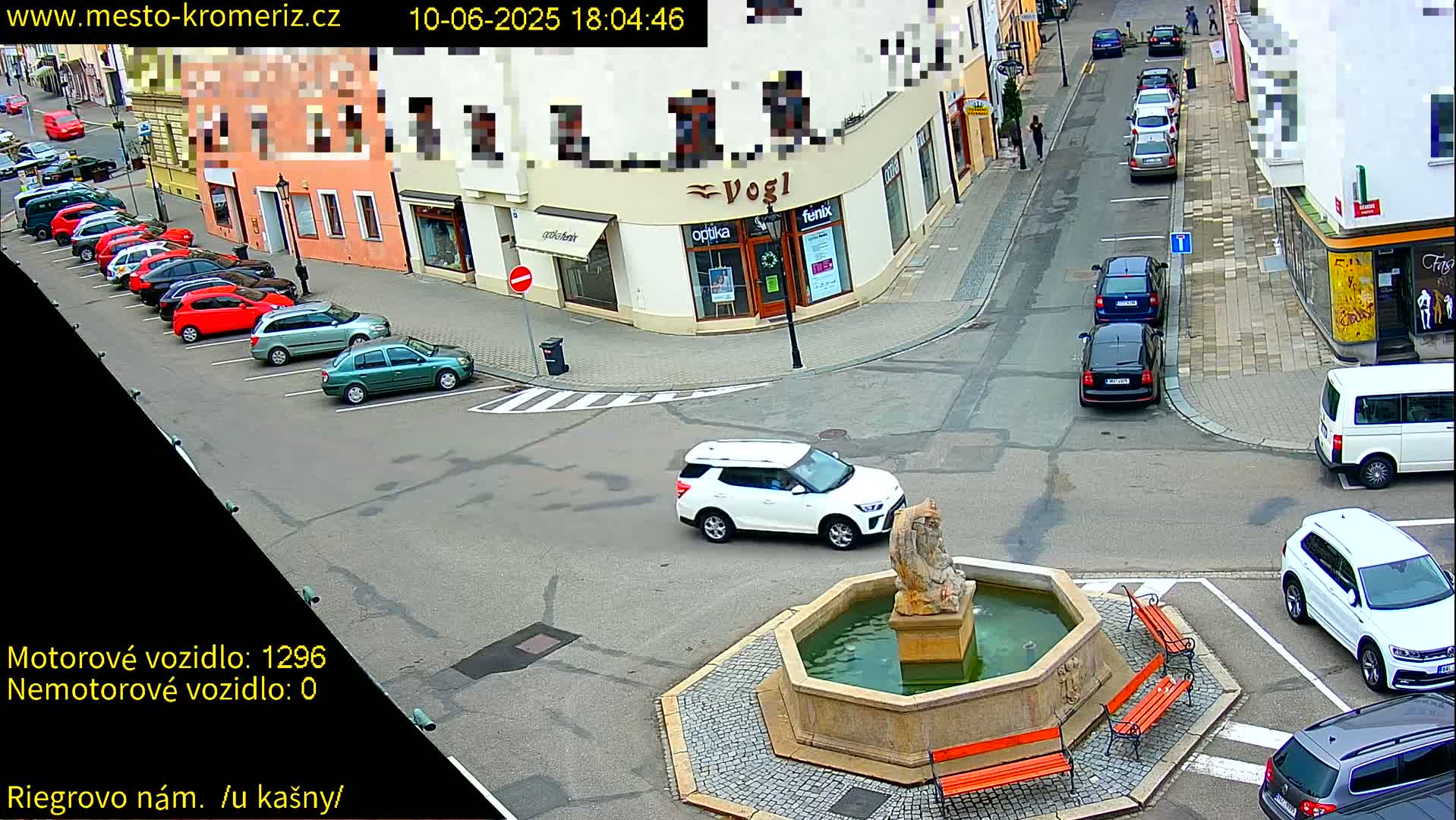 A town square with a fountain at its center, surrounded by parked cars and buildings, on a partly sunny day.