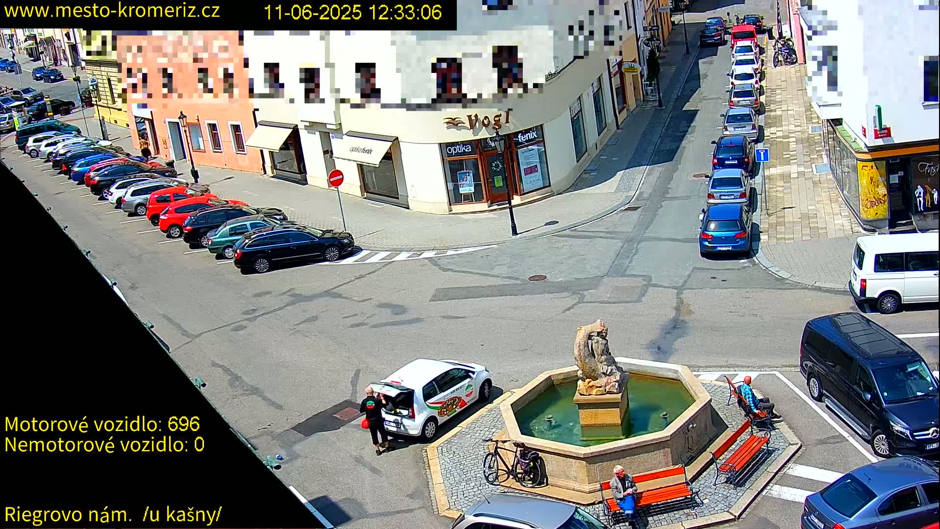 A sunny town square scene shows a fountain, parked cars, people sitting on benches, and a small white delivery vehicle.