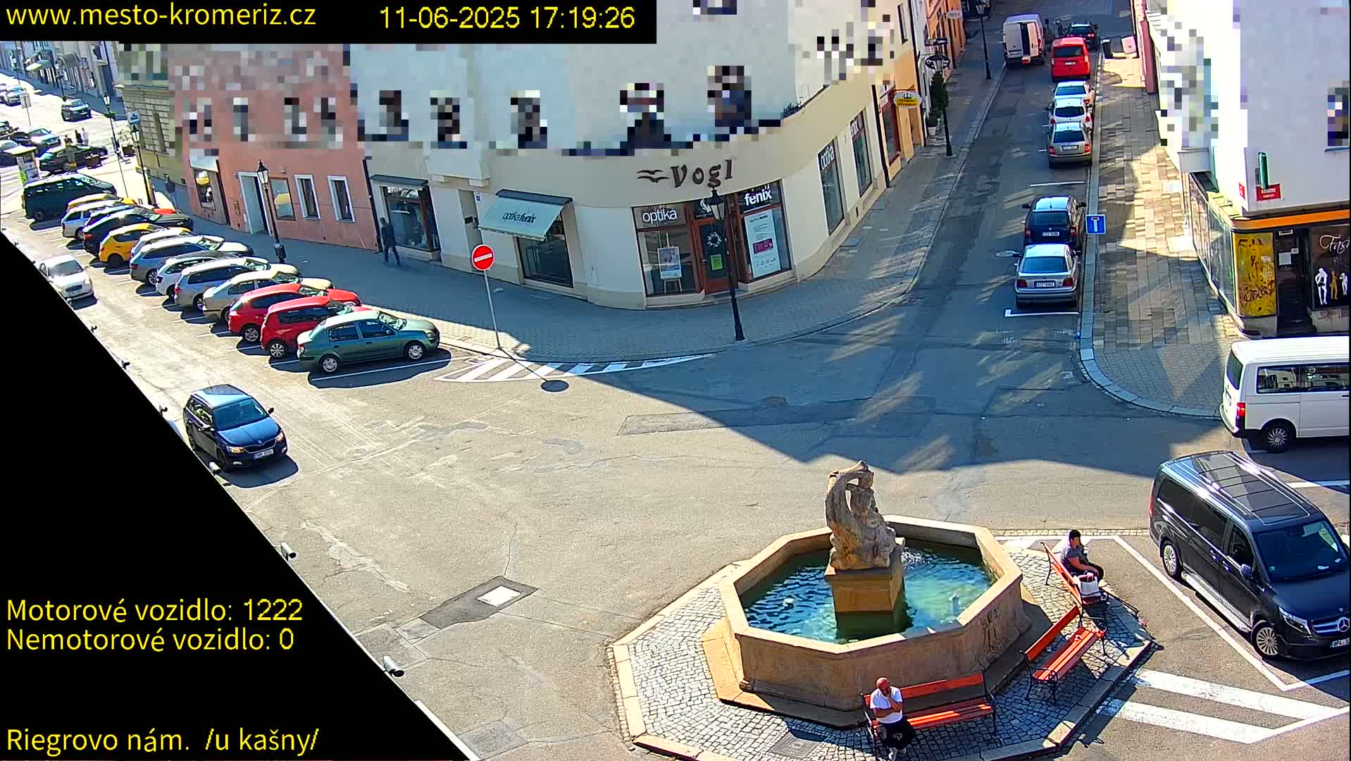 A sunny town square features a fountain, several parked cars, and a few pedestrians sitting on benches.
