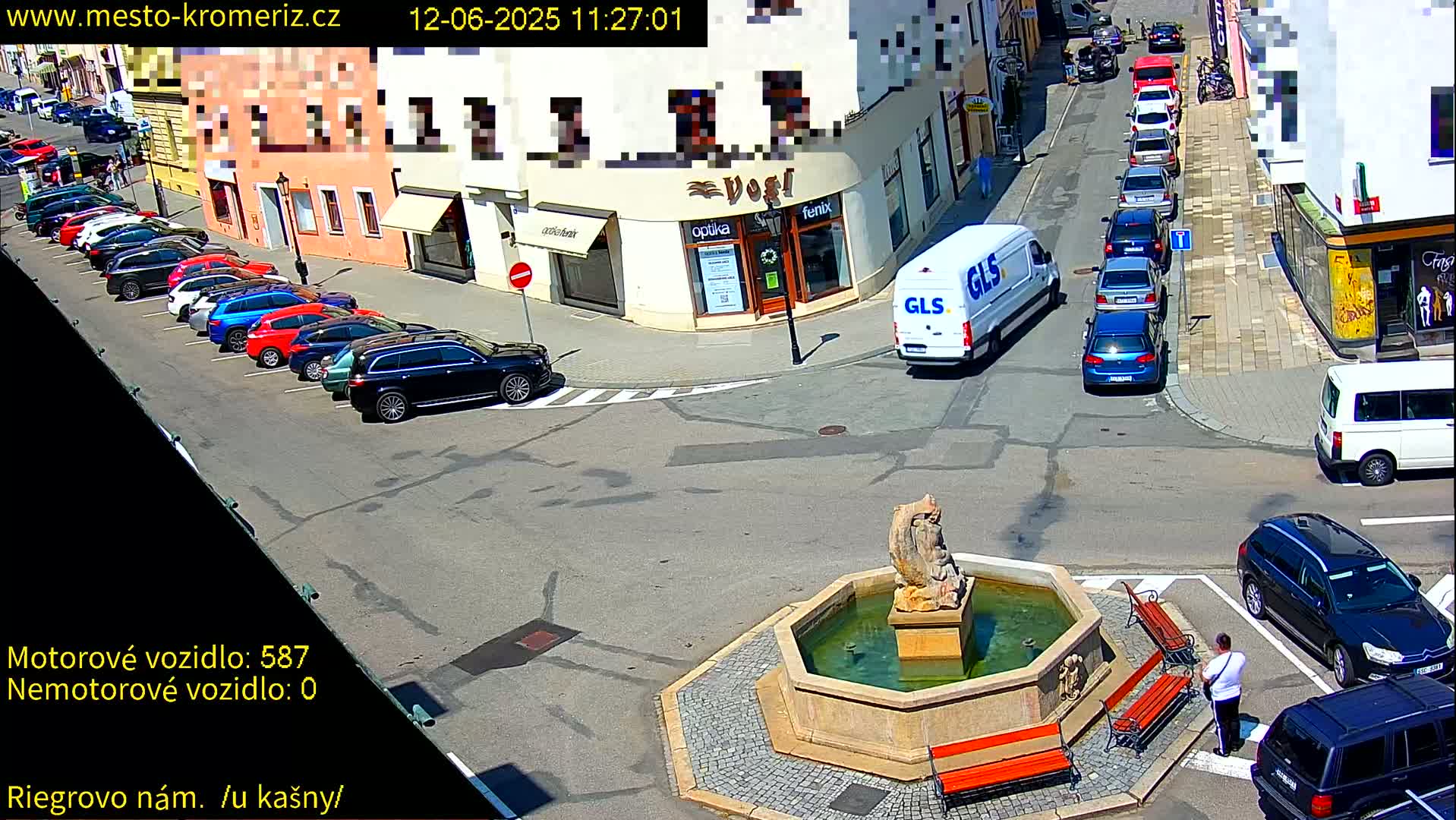 A sunny town square scene shows parked cars lining a street, a delivery van, a stone fountain with benches, and a person standing near the fountain.