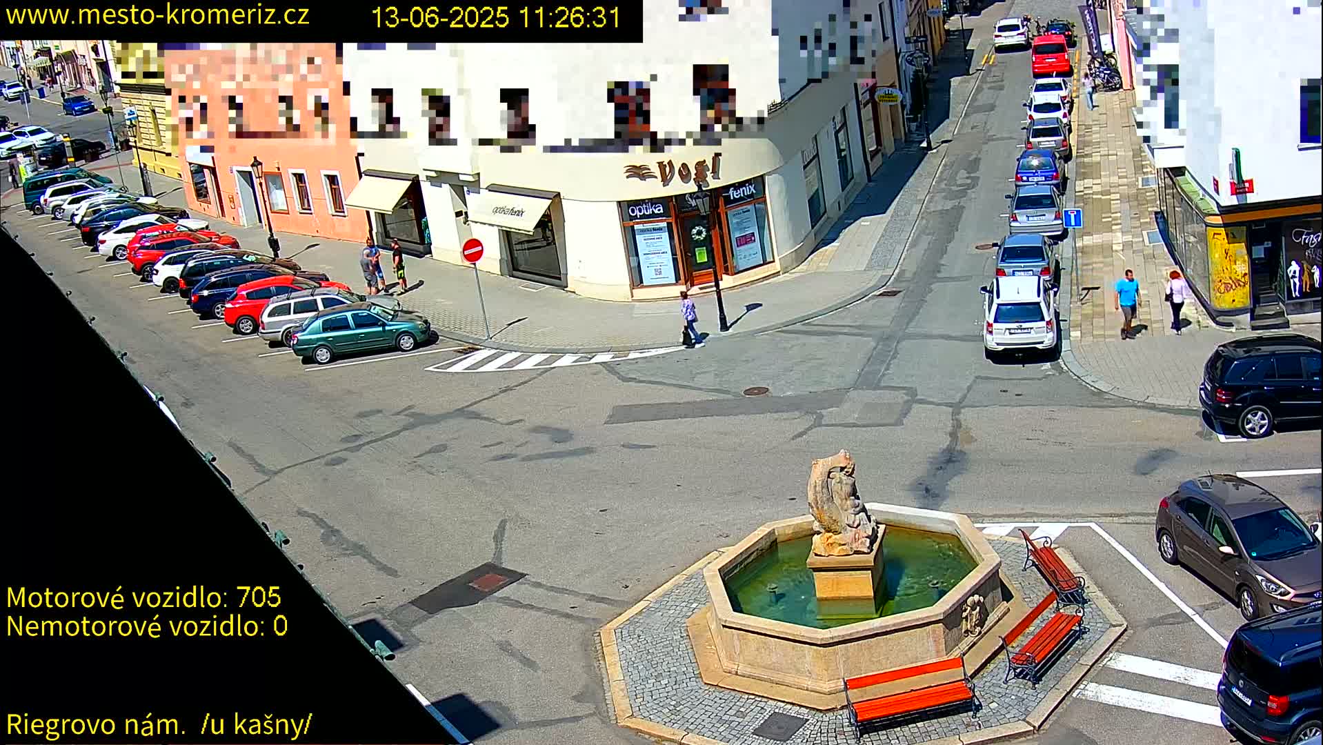 A sunny town square with a fountain, several parked cars, and a few pedestrians is viewed from above.