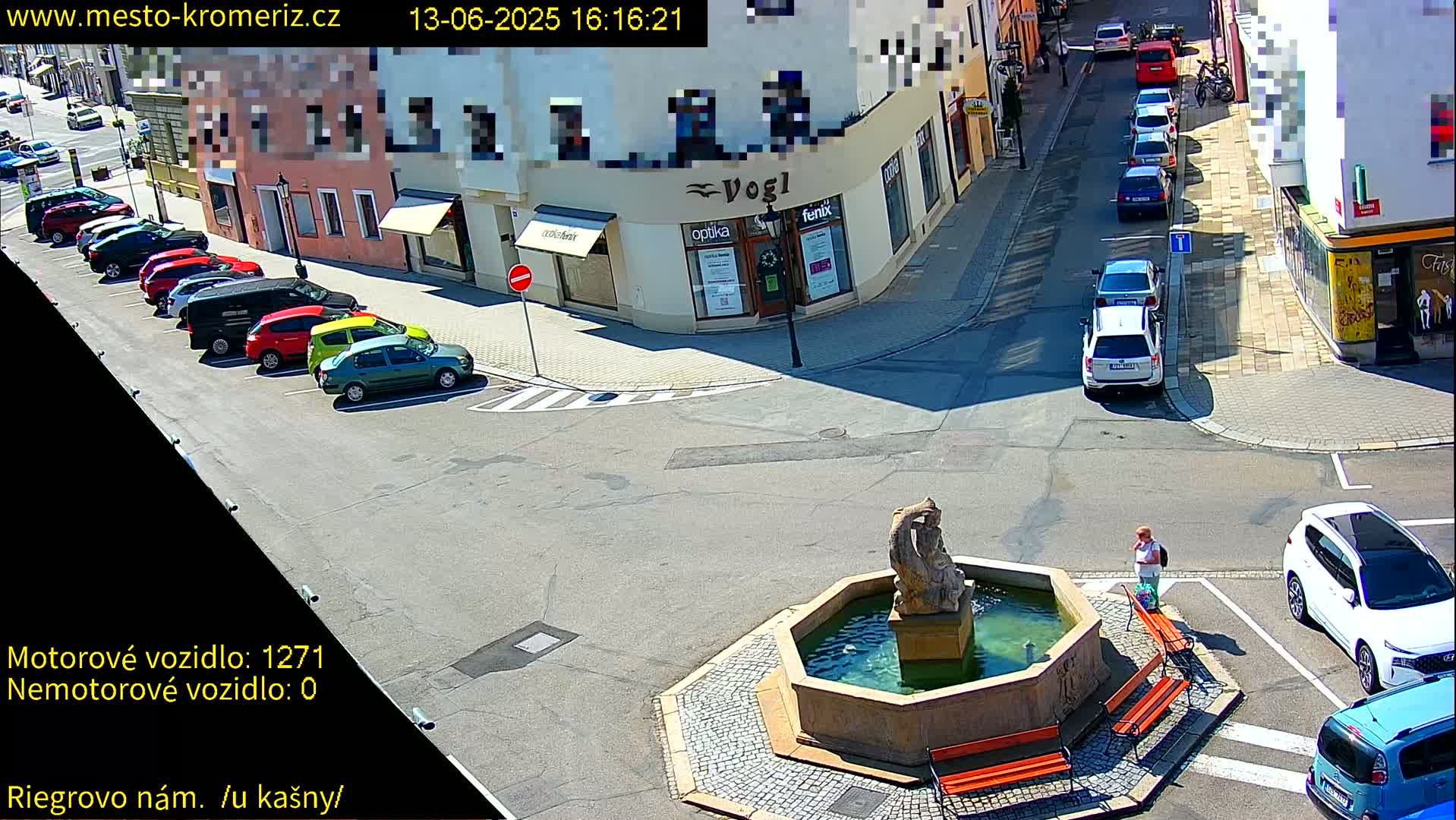 A sunny town square with a fountain, several parked cars, and a person standing near a bench.