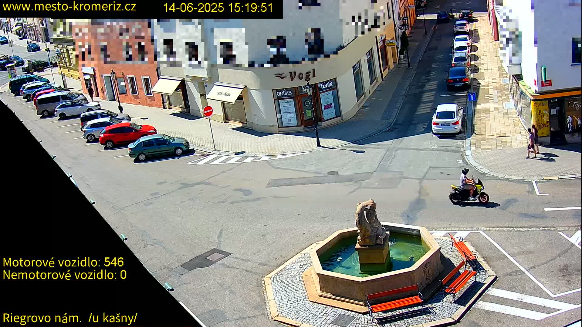 A sunny street scene shows a town square with a fountain, several parked cars, and a person riding a scooter.