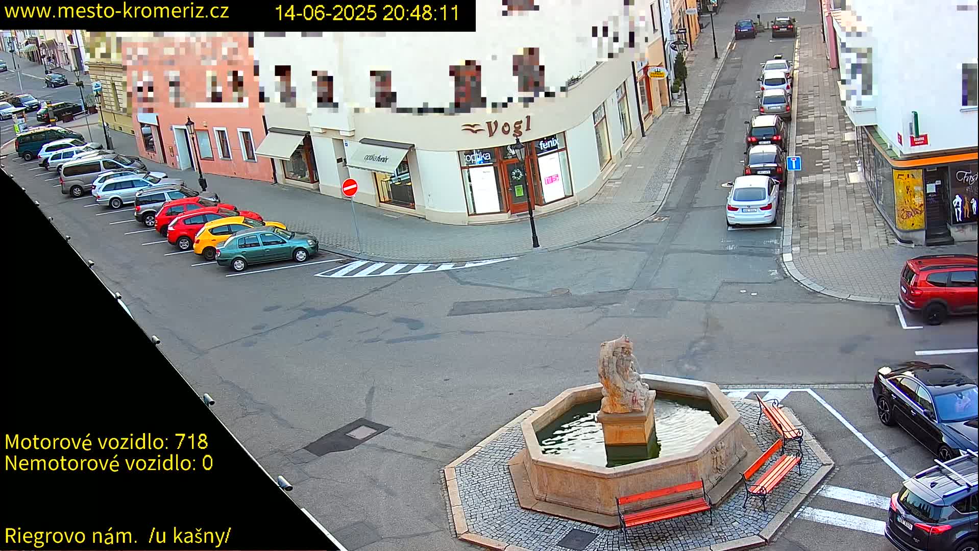 A sunny street scene shows a town square with a fountain, several parked cars, and a person riding a scooter.