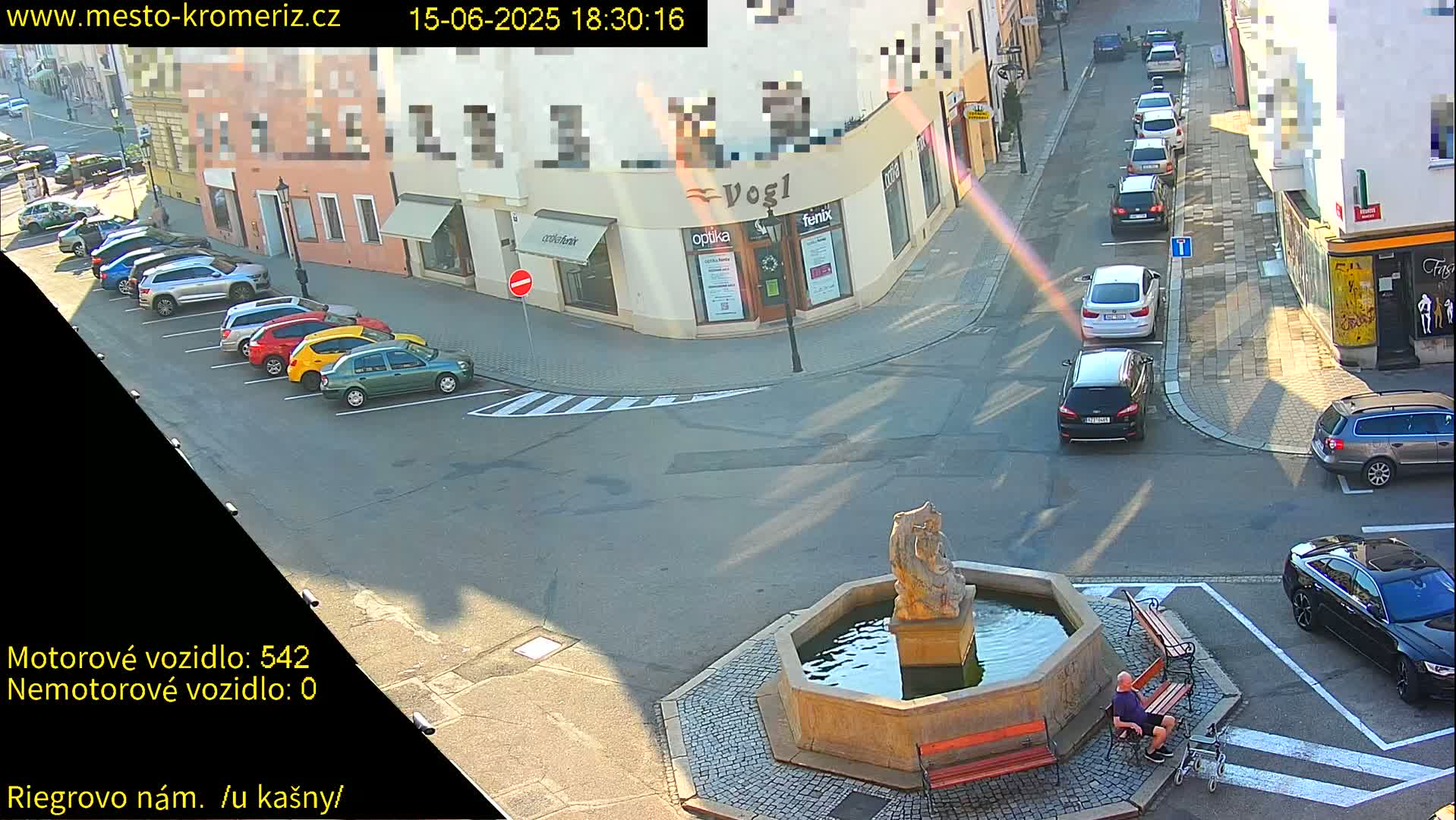 A sunny outdoor scene shows a town square with a fountain, several parked cars, and a person sitting on a bench.