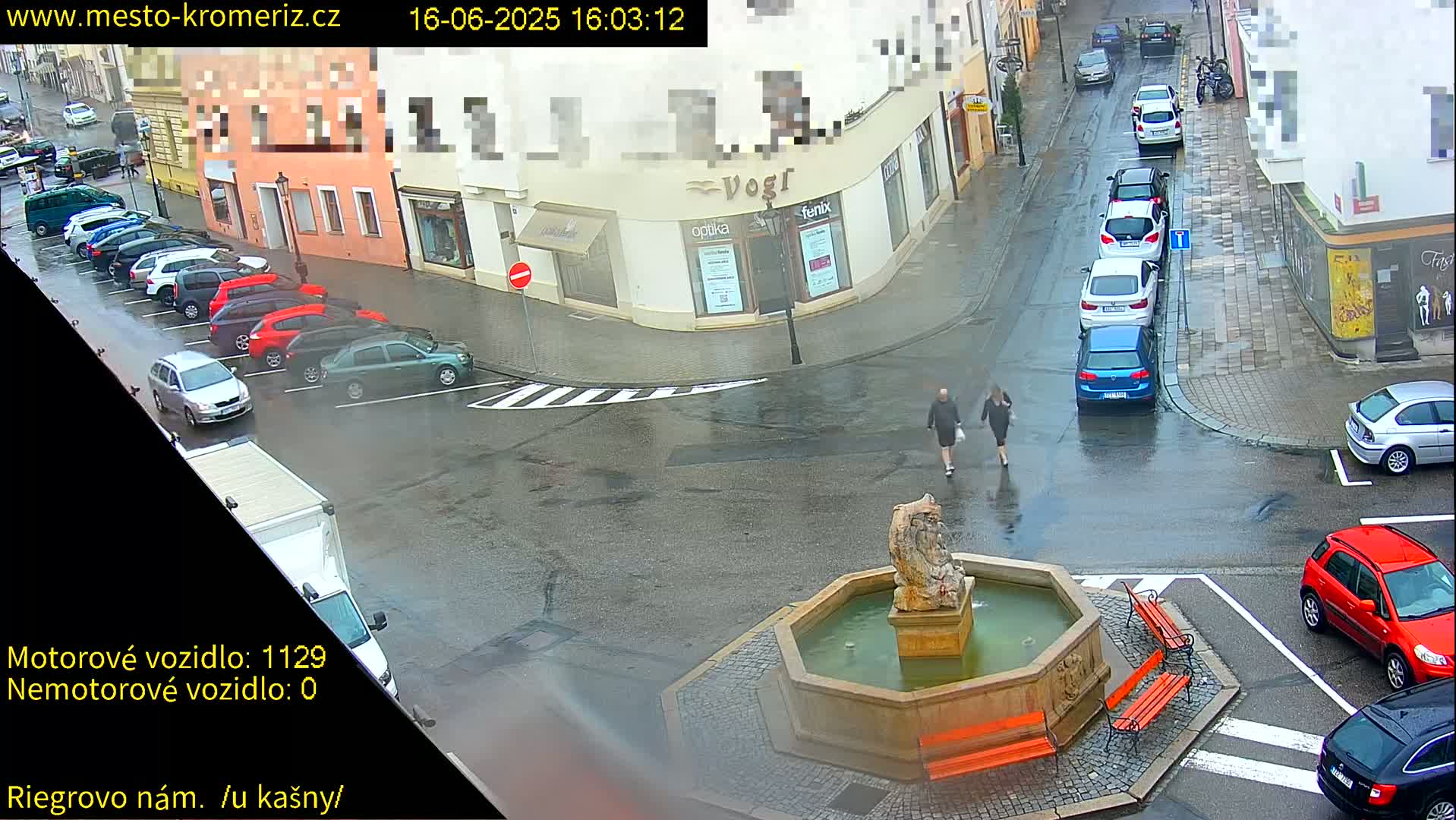 A town square with a fountain in the center is shown on a rainy day, with cars parked along the street and two pedestrians walking.