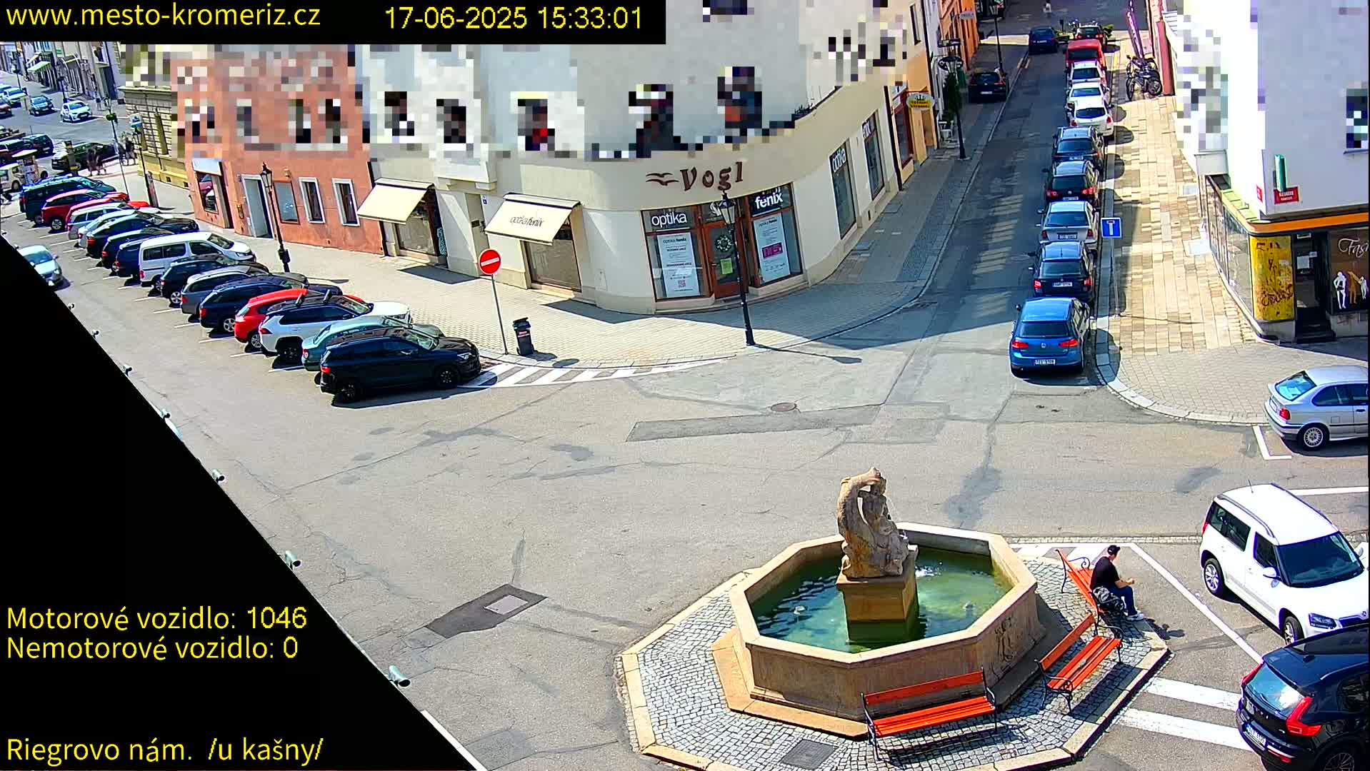 A sunny town square with a fountain, several parked cars, and a person sitting on a bench.