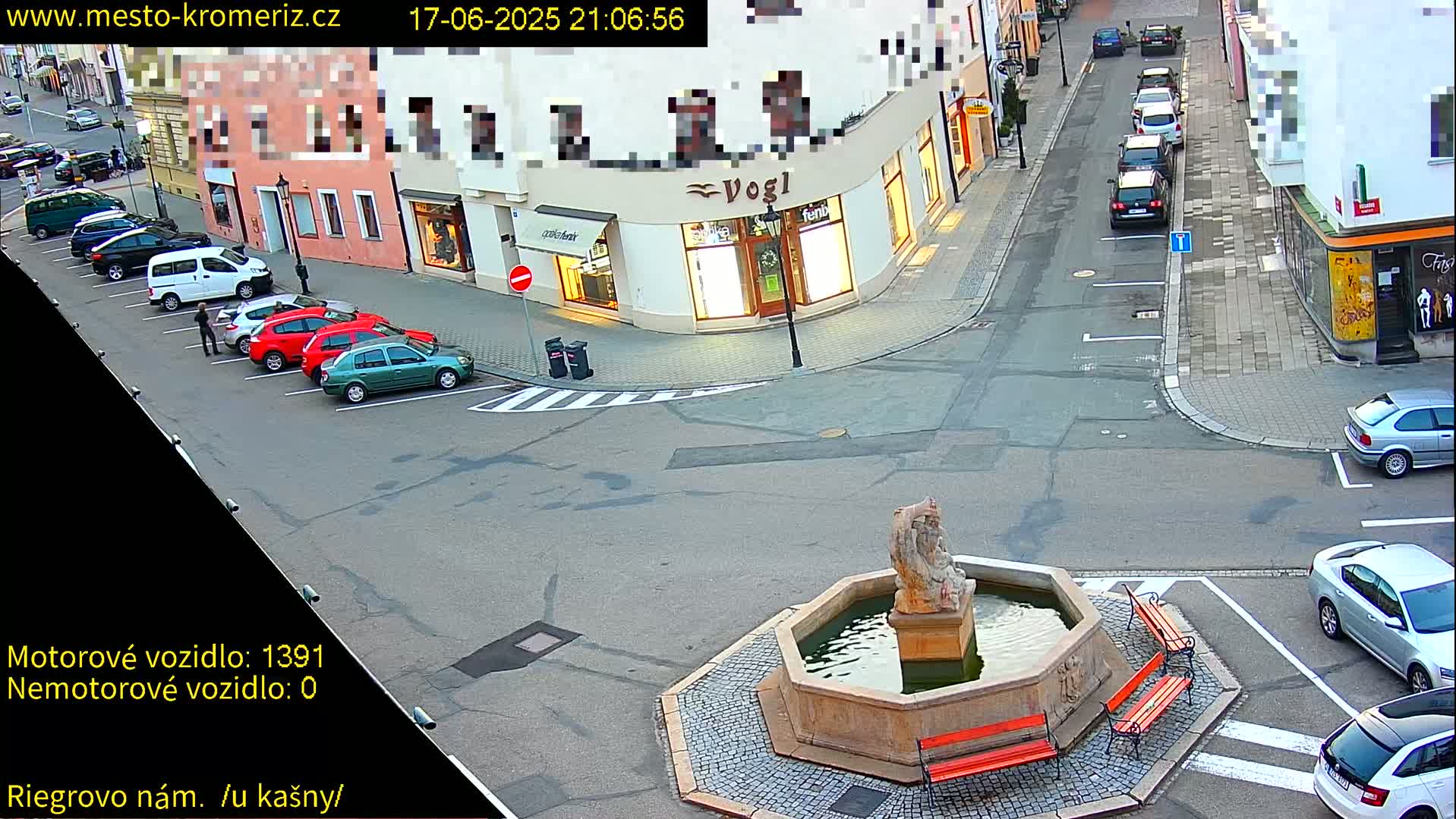 A nighttime street scene shows several parked cars surrounding a stone fountain with benches, in a town square.