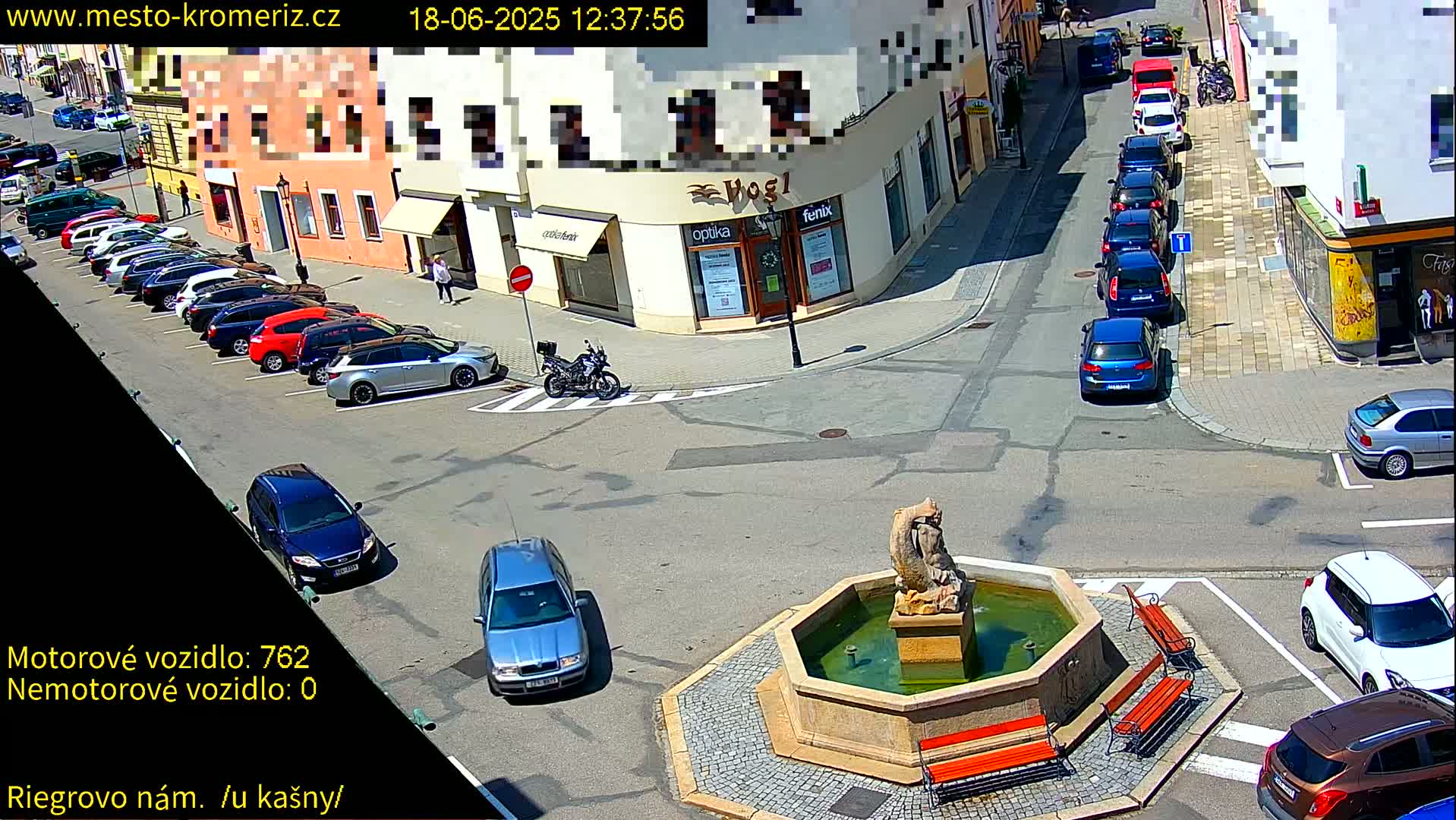 A sunny town square with a fountain, several parked cars, and a motorcycle, surrounded by buildings.