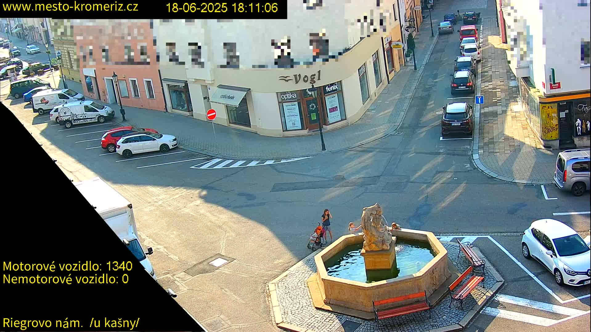 A sunny town square features a stone fountain surrounded by benches, with several parked cars visible nearby and a few pedestrians near the fountain.