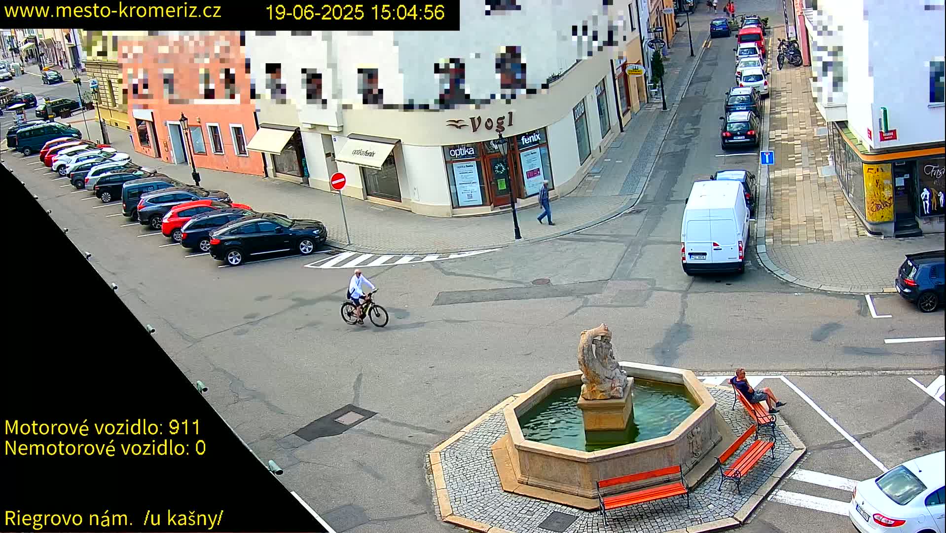 A town square with a fountain, several parked cars, a person biking, and a person sitting on a bench, under sunny conditions.