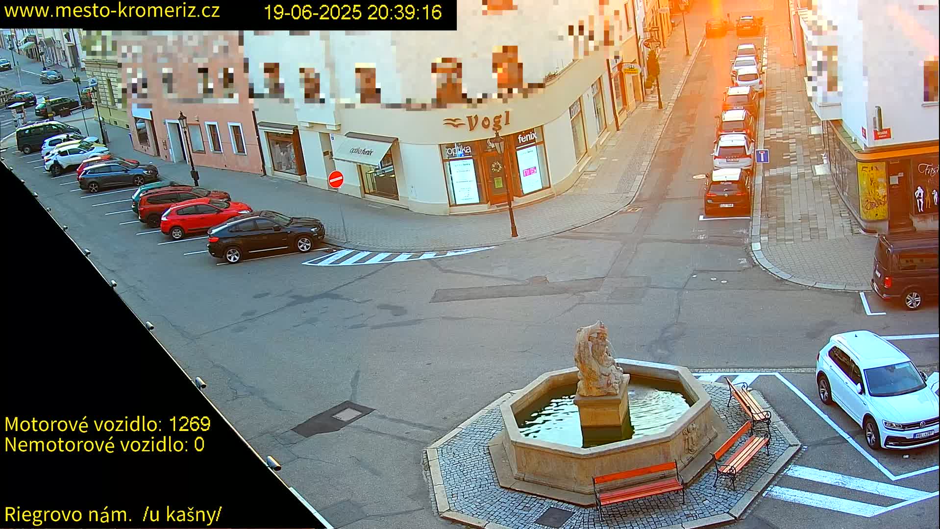 A street scene at sunset shows several cars parked along the side of a street, a fountain in the center of a plaza, and more cars parked in the plaza, with several buildings in the background.