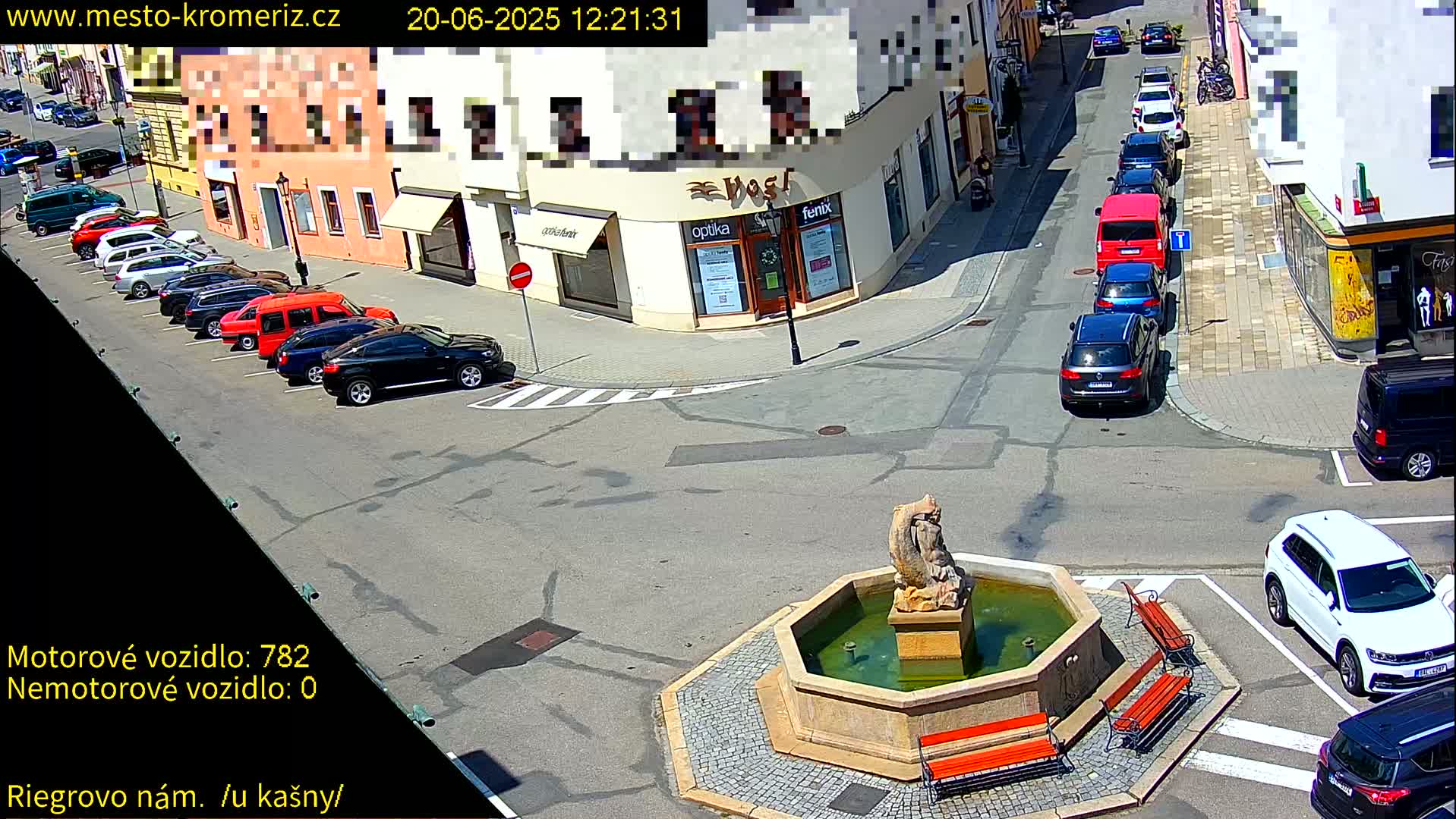 A sunny town square with a stone fountain and several parked cars, some cars are also driving on the street.